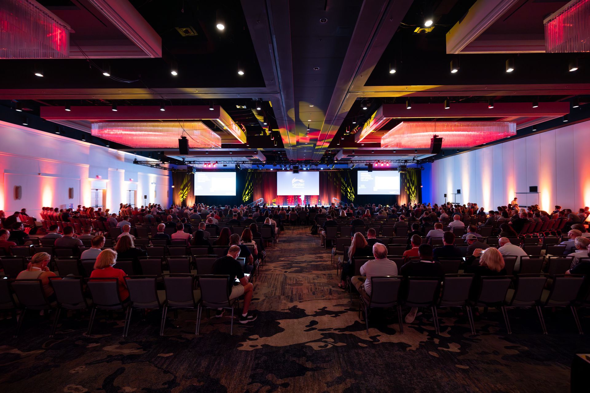 Large conference hall, filled audience facing a stage with screens, brightly lit in red and white.