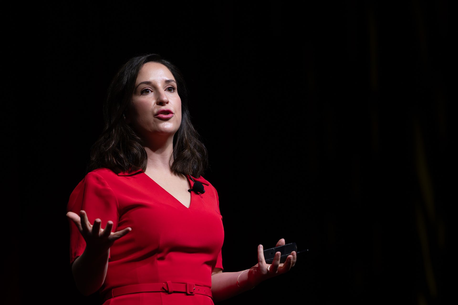 Woman in red dress speaking on stage, gesturing with hands, dark background.