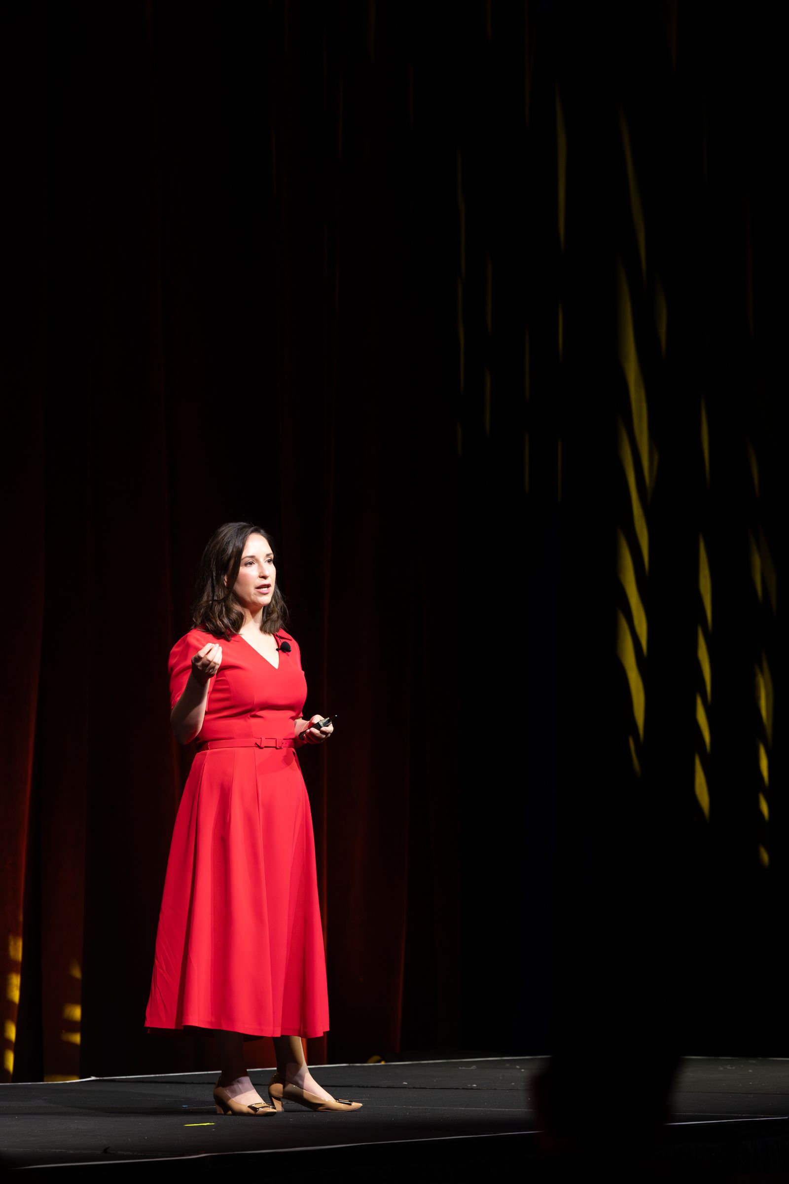 Woman in red dress speaking on stage, gesturing, with dark curtains and spotlight.