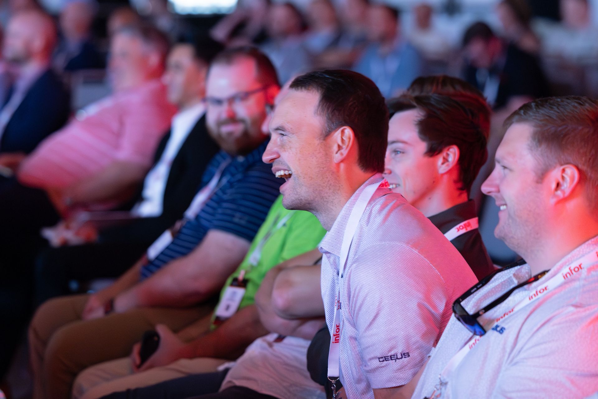 Audience in an event, laughing. People seated in rows, illuminated by pink and white light.