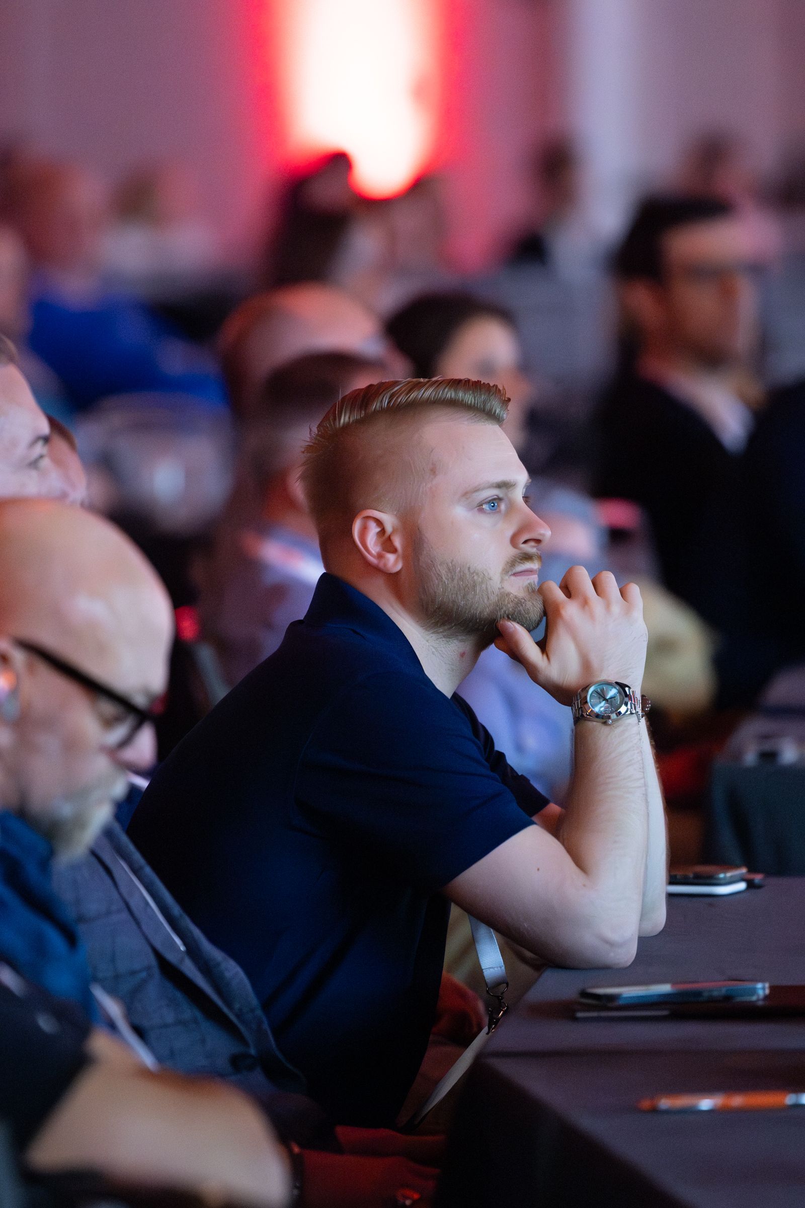 Man in blue shirt, hands clasped under chin, at a conference.