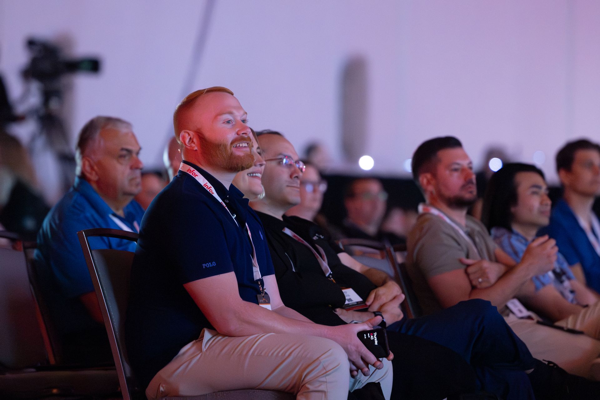 Audience members seated at an event, some smiling, looking at the presentation, indoors, with soft lighting.