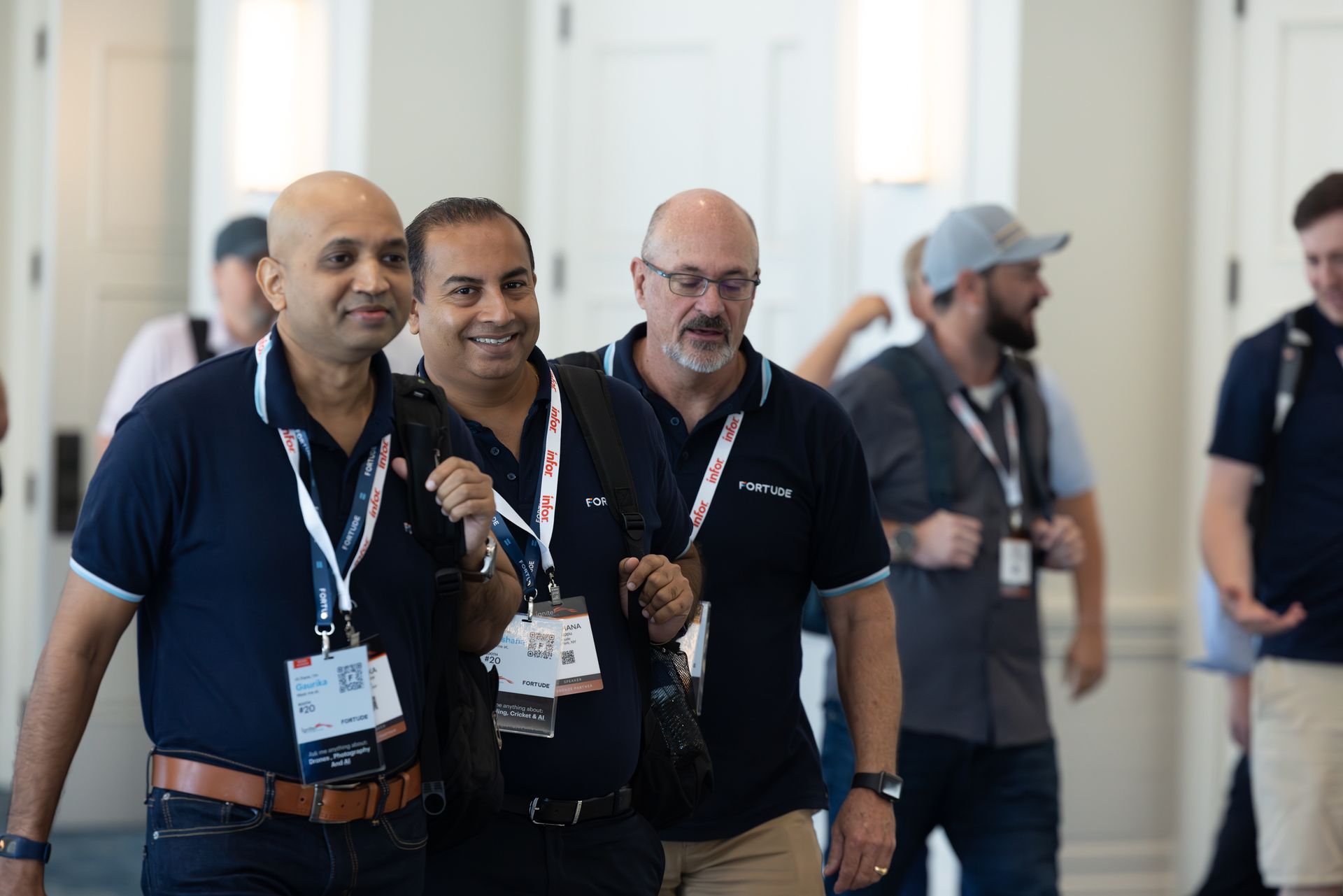 Three men in blue shirts walk and smile, wearing lanyards and carrying bags, in a hallway.
