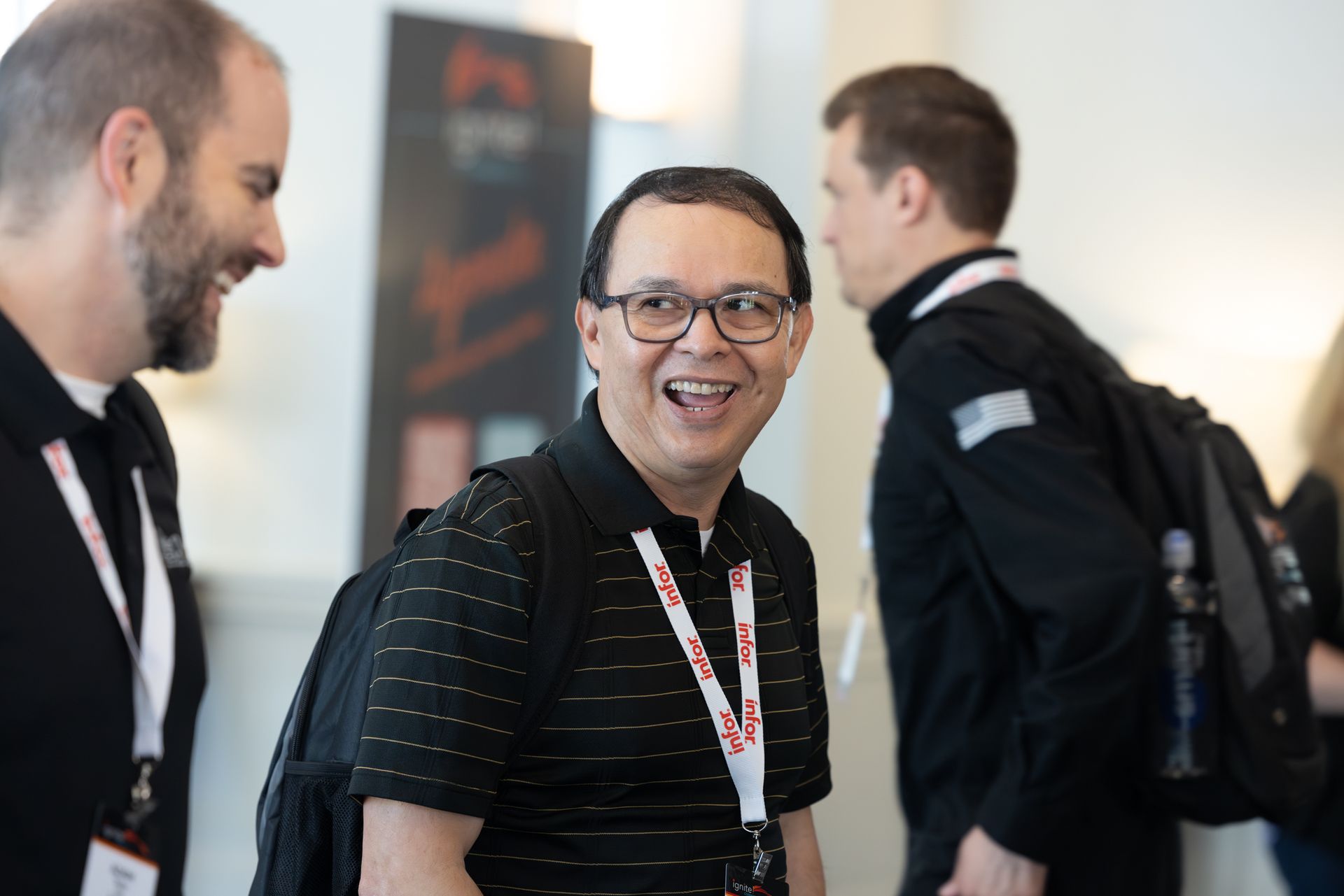 Man with glasses smiles, wearing black shirt and backpack, talking with others indoors.