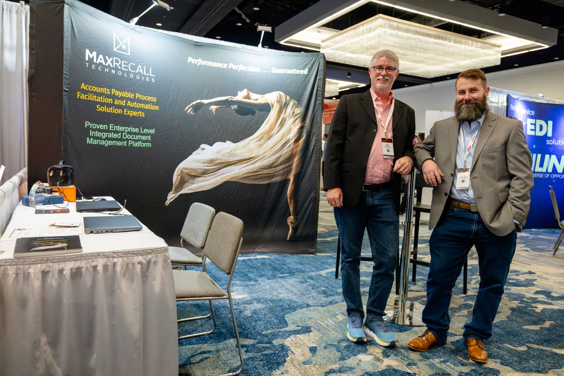 Two men stand at a trade show booth with a banner. One wears a blazer and jeans, the other a sport coat and jeans.