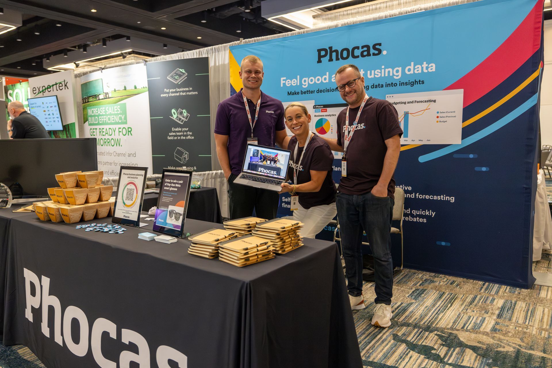 Three people at a trade show booth promoting Phocas, smiling and holding a laptop, blue and black booth.