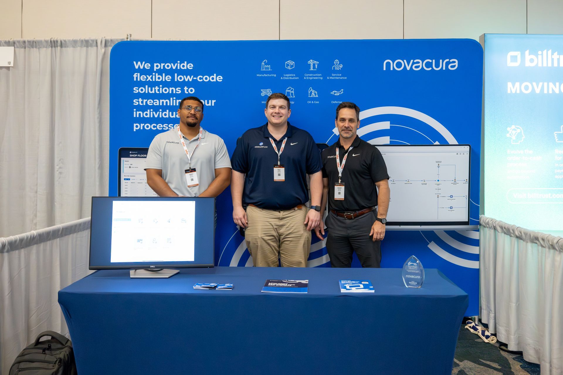 Three men at a Novacura trade show booth; blue backdrop, table with a computer screen, and a blue tablecloth.