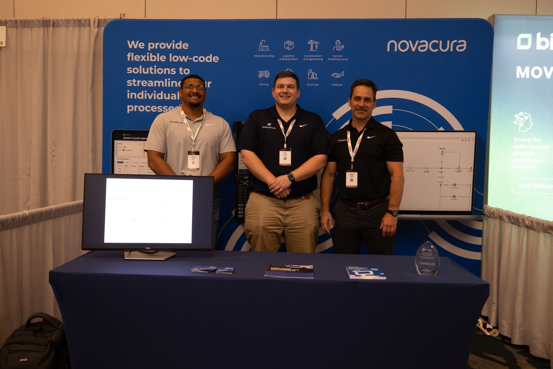 Three men at a trade show booth with a computer monitor, Novacura banner, and a table with brochures.