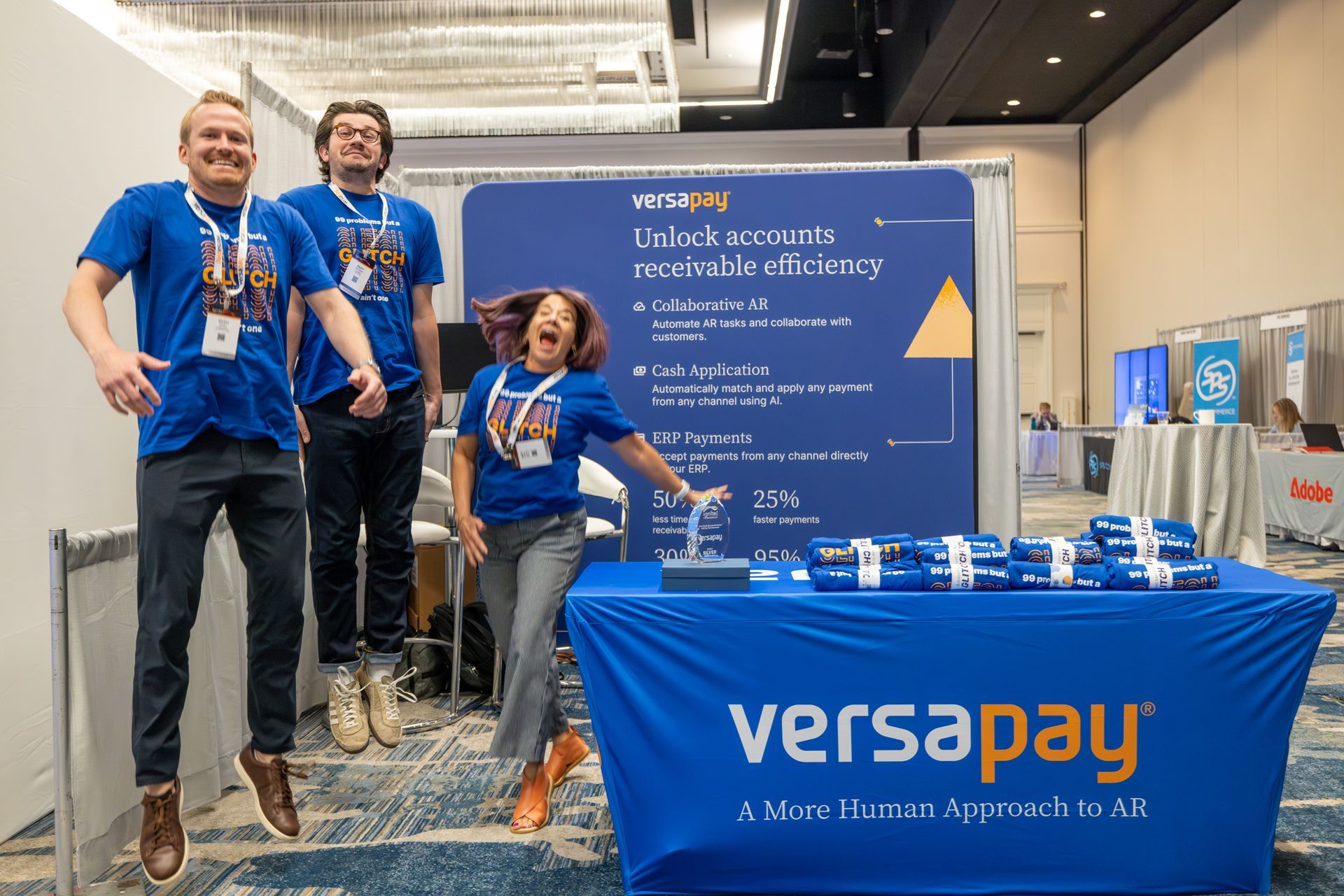 Three people in blue shirts jump for joy at the Versapay booth at a conference.