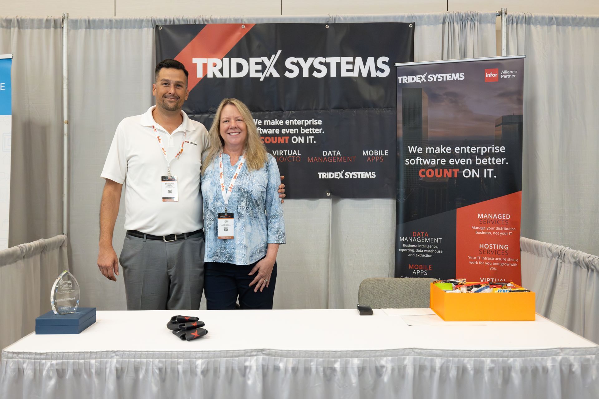 Two people stand behind a TRIDEX SYSTEMS booth at a trade show.
