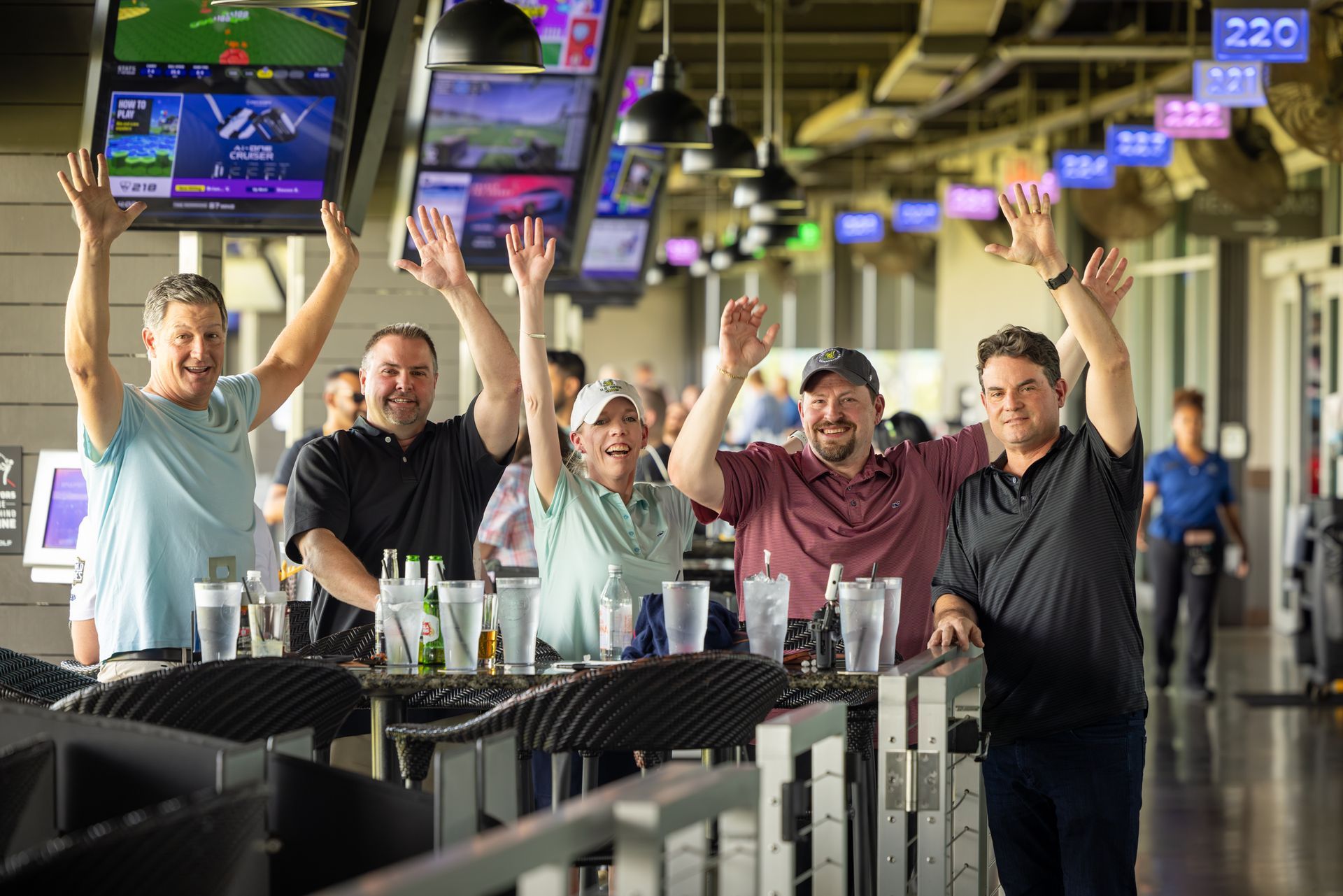 Group of people cheering at Topgolf, arms raised. Drinks on the table, screens overhead.