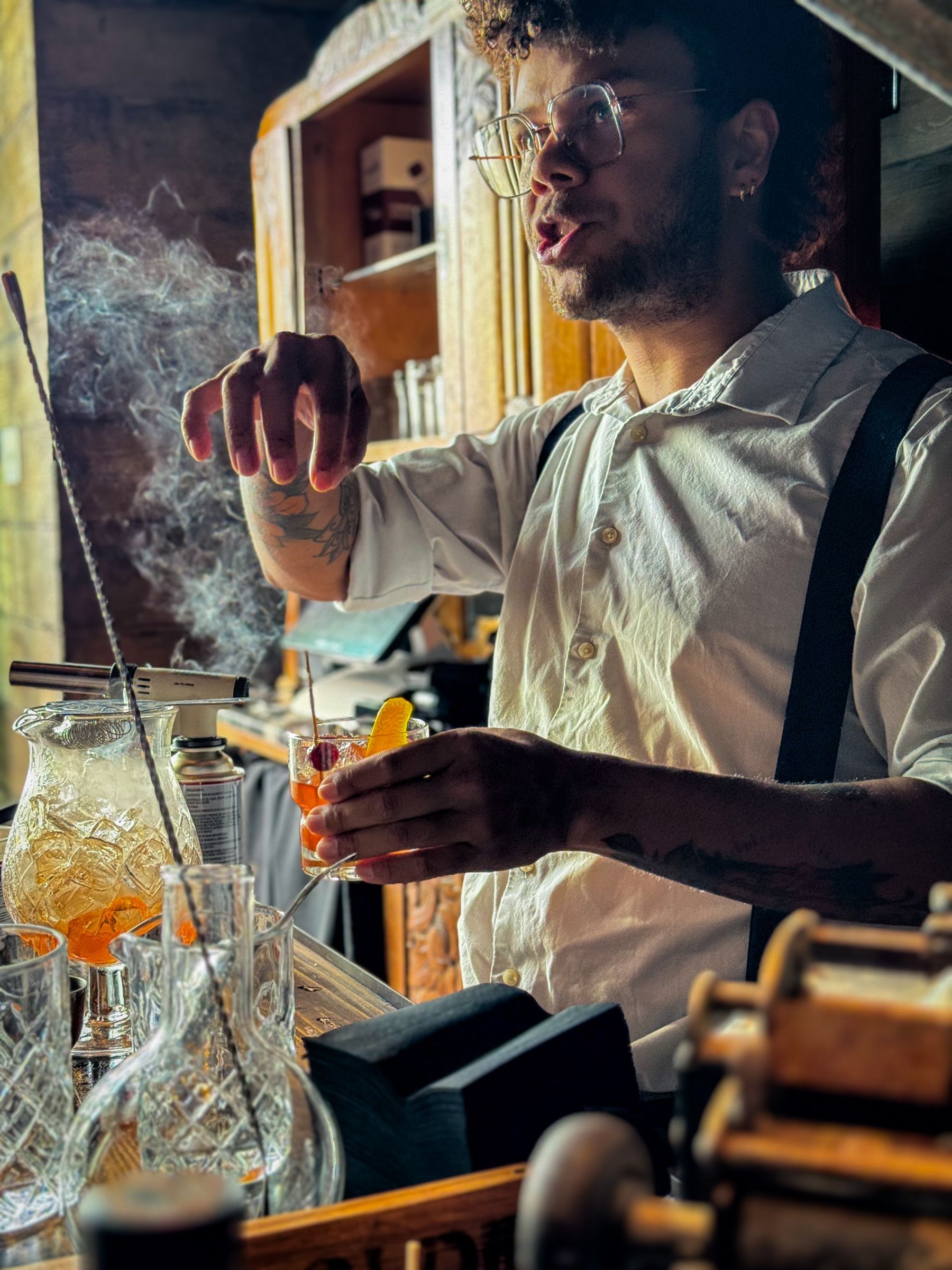 Bartender with curly hair makes a cocktail, smoke rising, wearing suspenders.