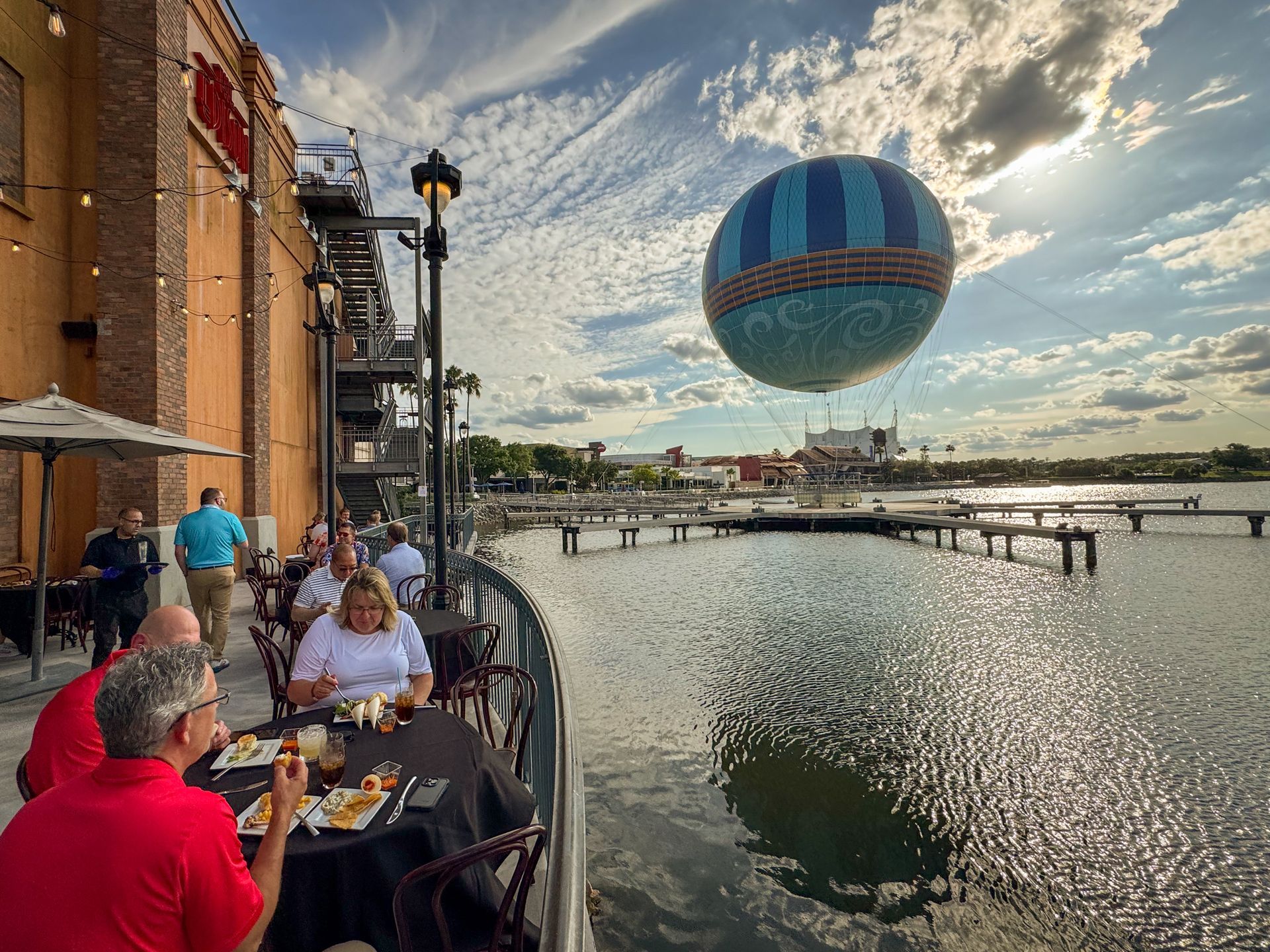 Outdoor waterfront dining with balloon, building. People eating at tables under umbrellas. Sunny day.