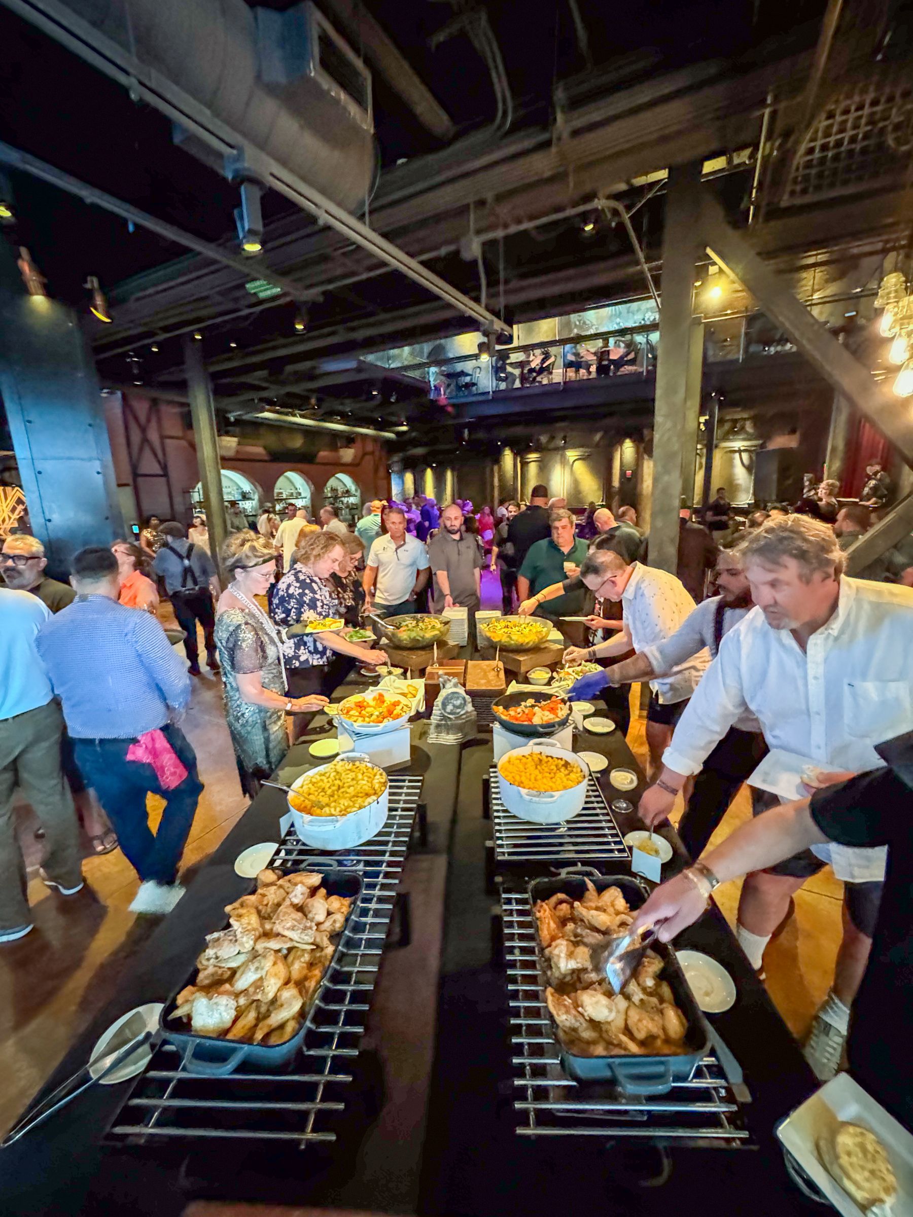 People at a buffet with multiple food selections. Inside, dark room, many guests, food servers.