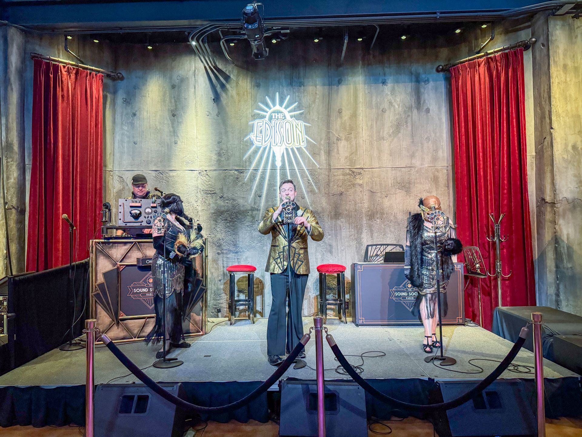Band on stage with brass instruments, wearing gold and unique outfits, red curtains, stage lights.