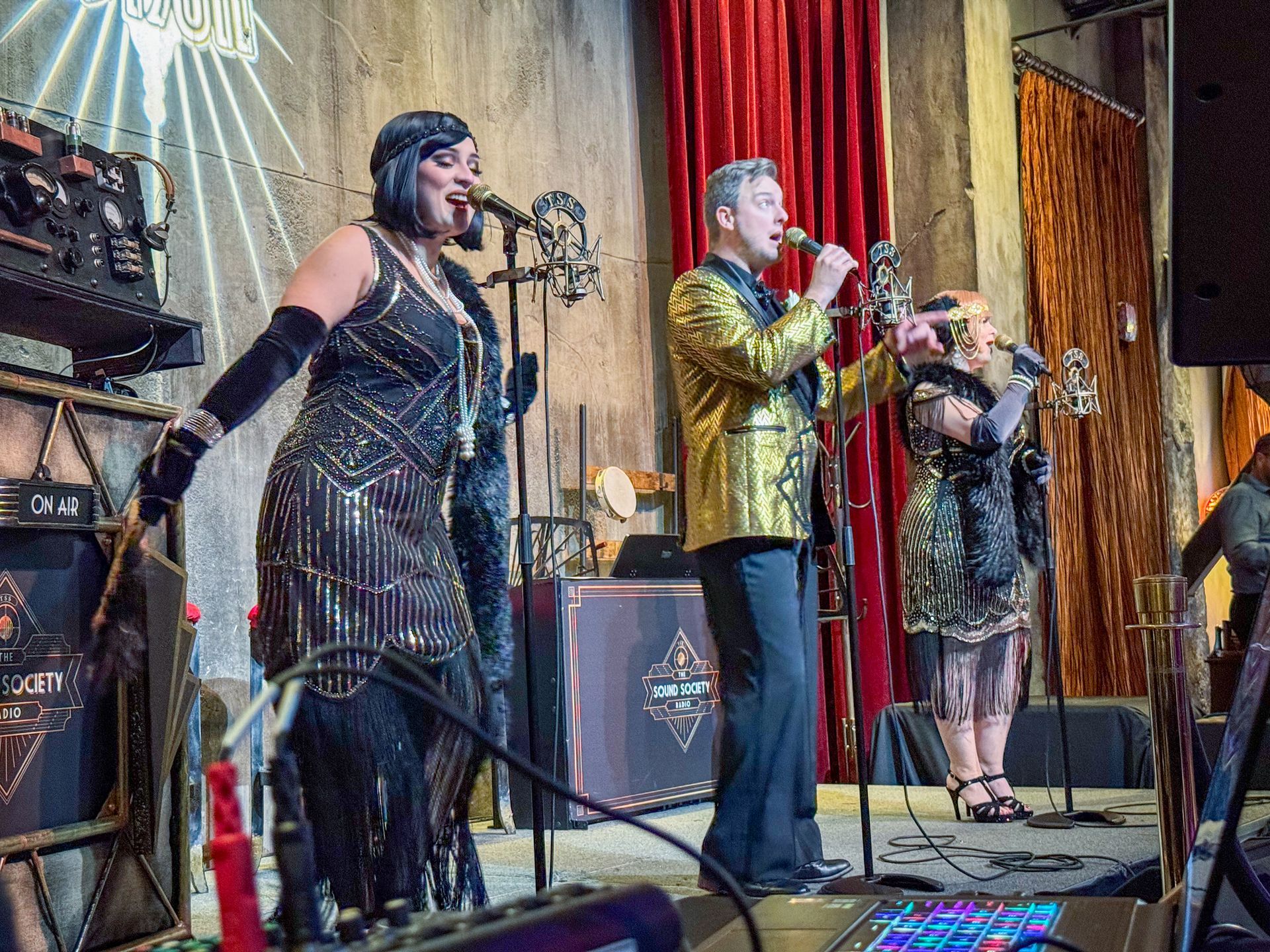 Band performing on stage, dressed in 1920s attire. Performers singing into microphones, stage backdrop.
