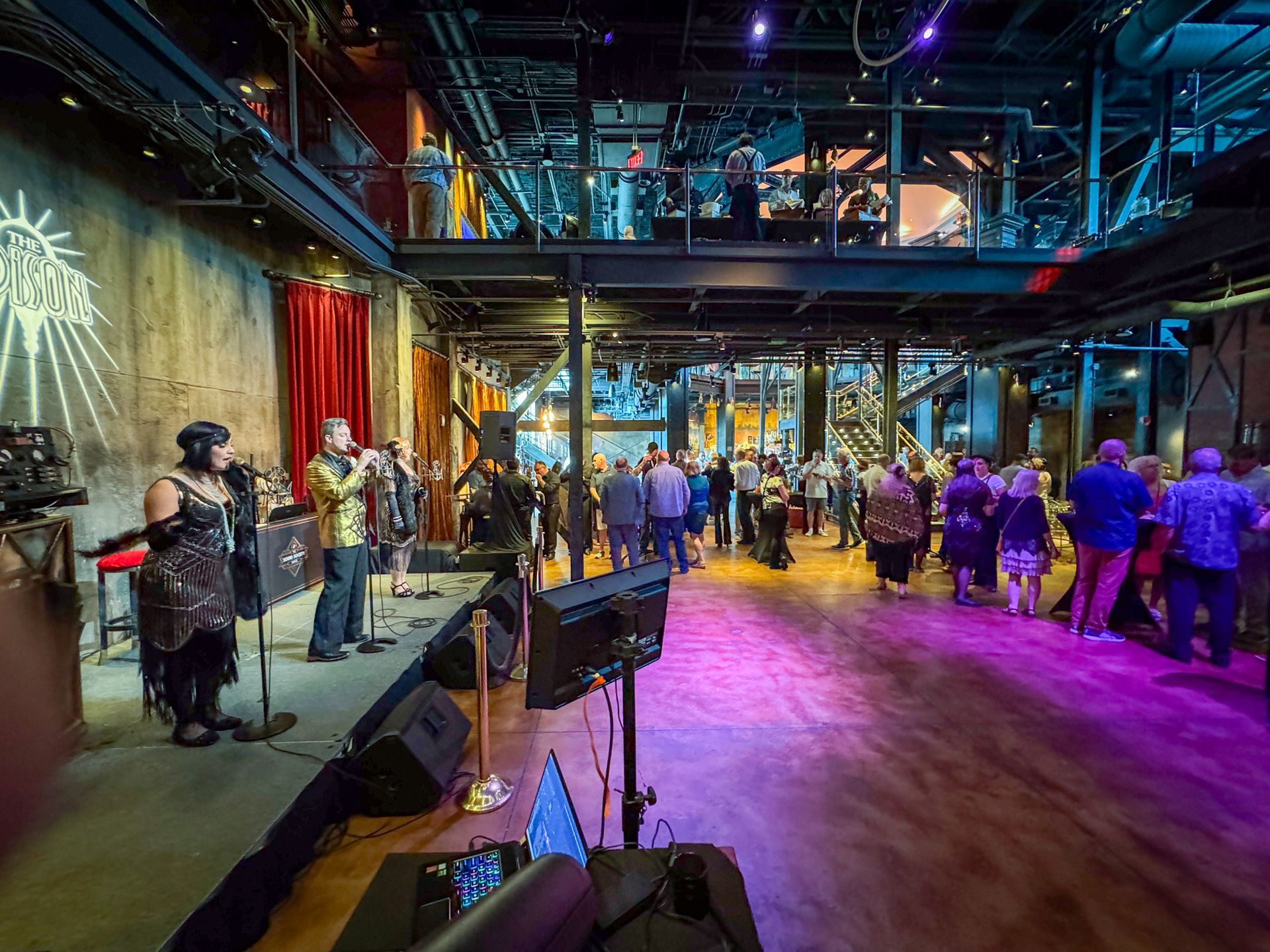 Band performing on stage in a large, industrial-style venue. Guests mingle, upper level viewing.