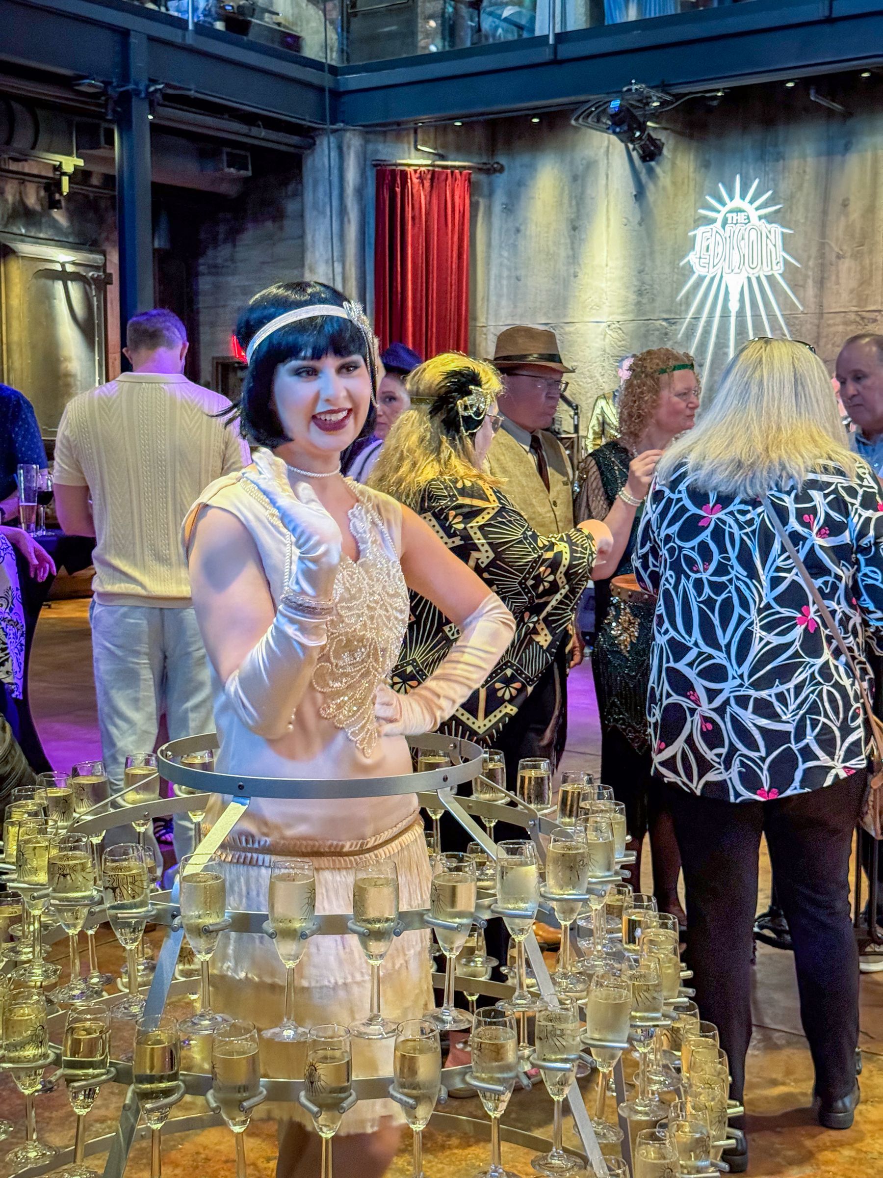Woman in flapper dress smiles near a champagne tower at a party.