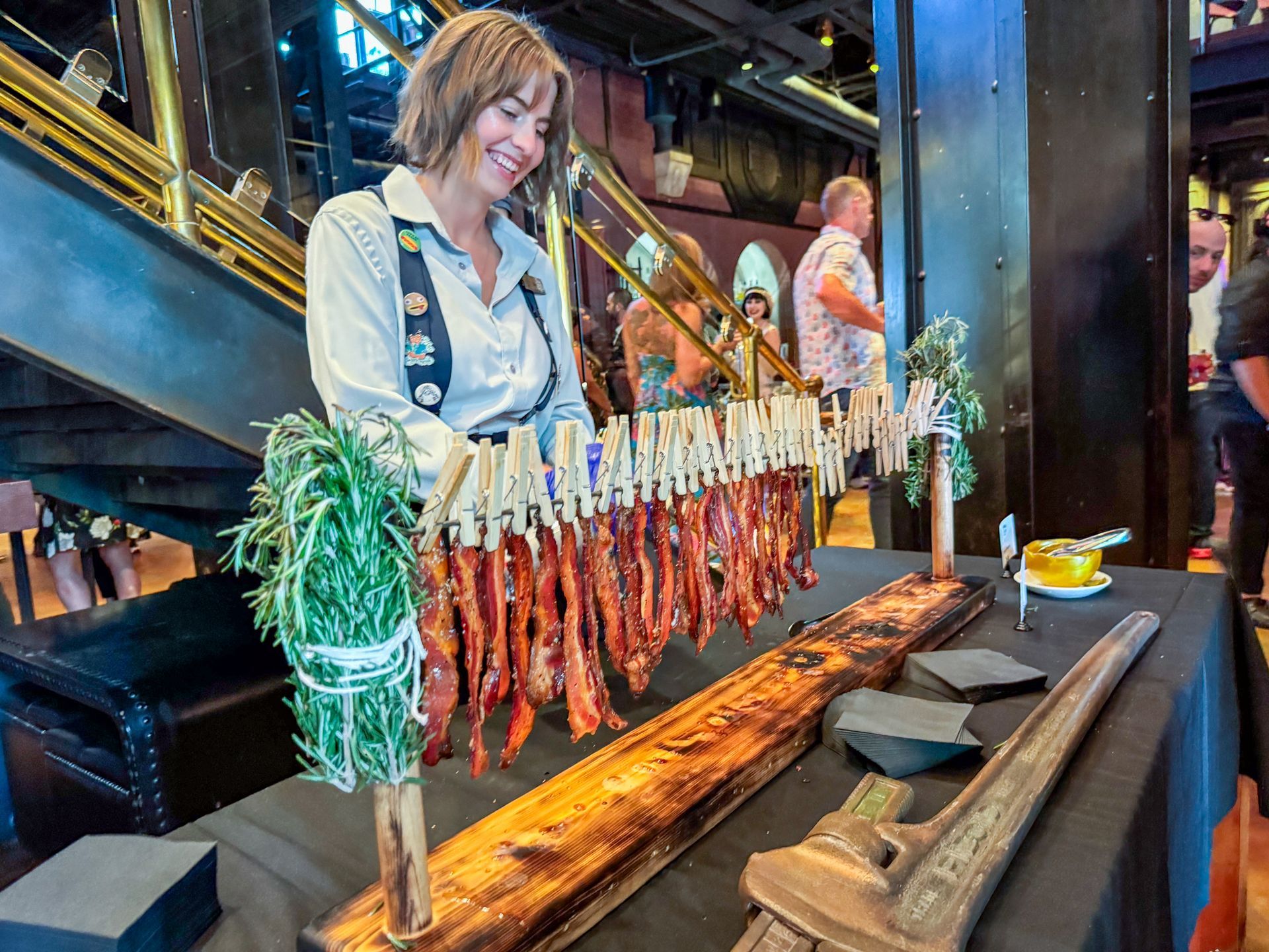 Woman smiling, displaying strips of bacon clipped to a wooden board, with rosemary, in a restaurant.
