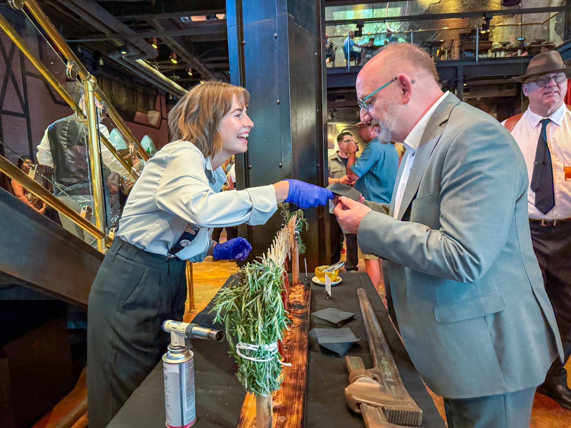 Woman in uniform offers food to a man in a suit. They stand at a wooden counter indoors; many people nearby.