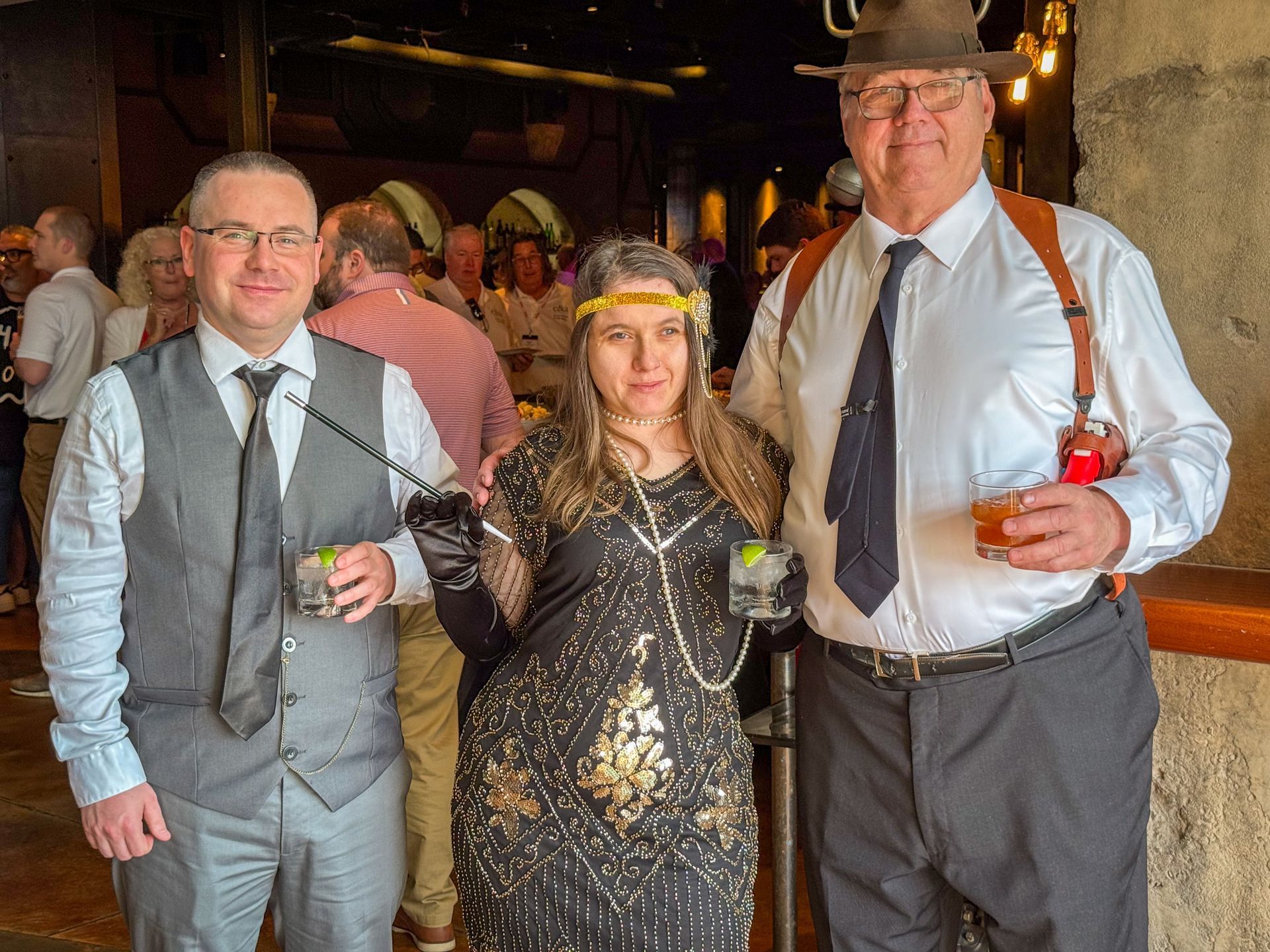 Three people in vintage attire pose, holding drinks, inside a dimly lit venue.