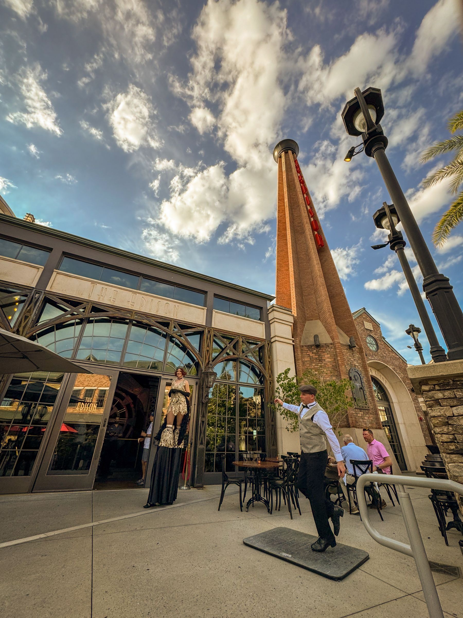 Exterior of a restaurant with a tall brick smokestack. Two employees gesture to the entrance; blue sky with clouds.