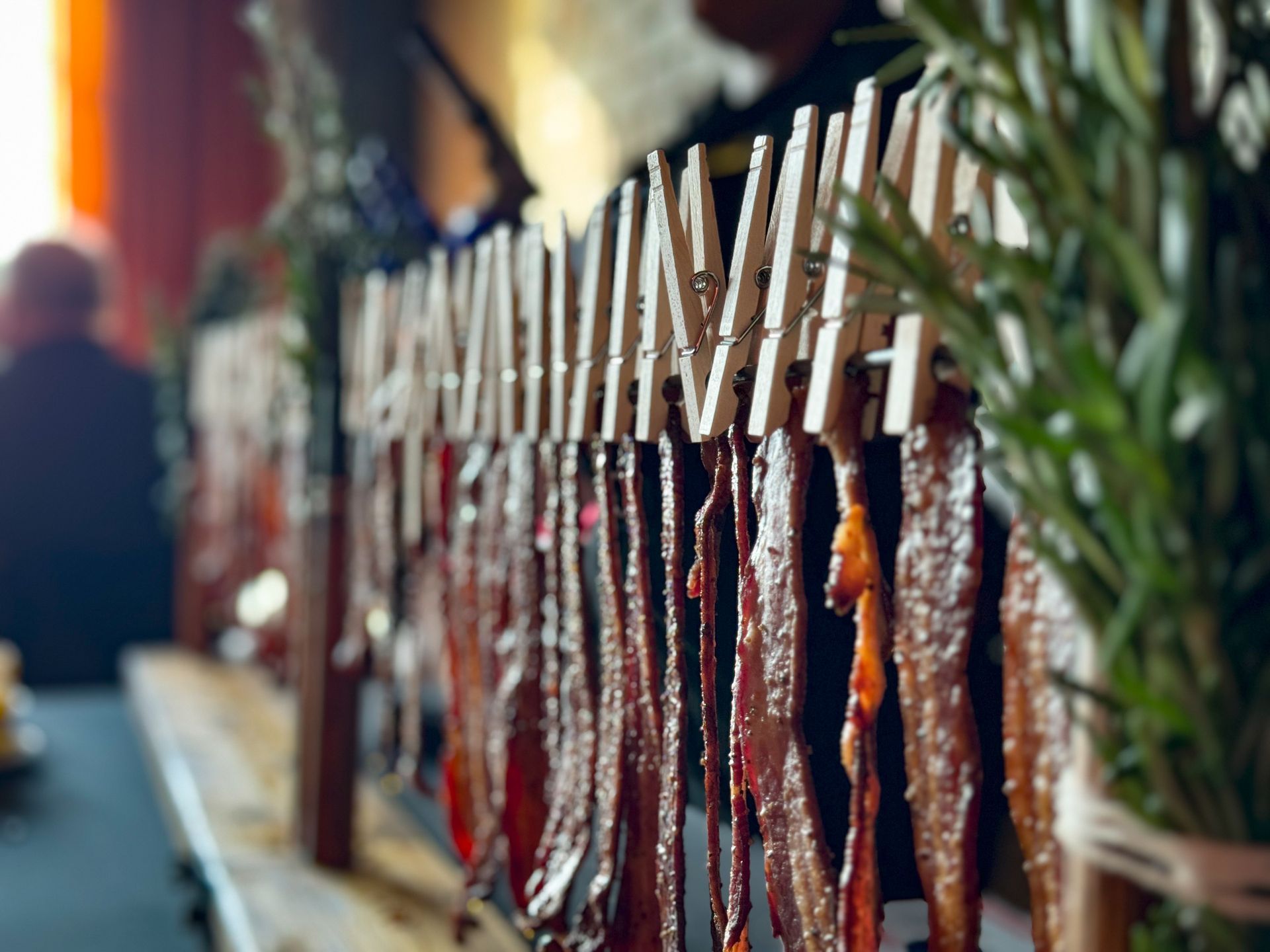 Strips of bacon clipped to clothespins on a wooden rack, rosemary sprigs in the background.