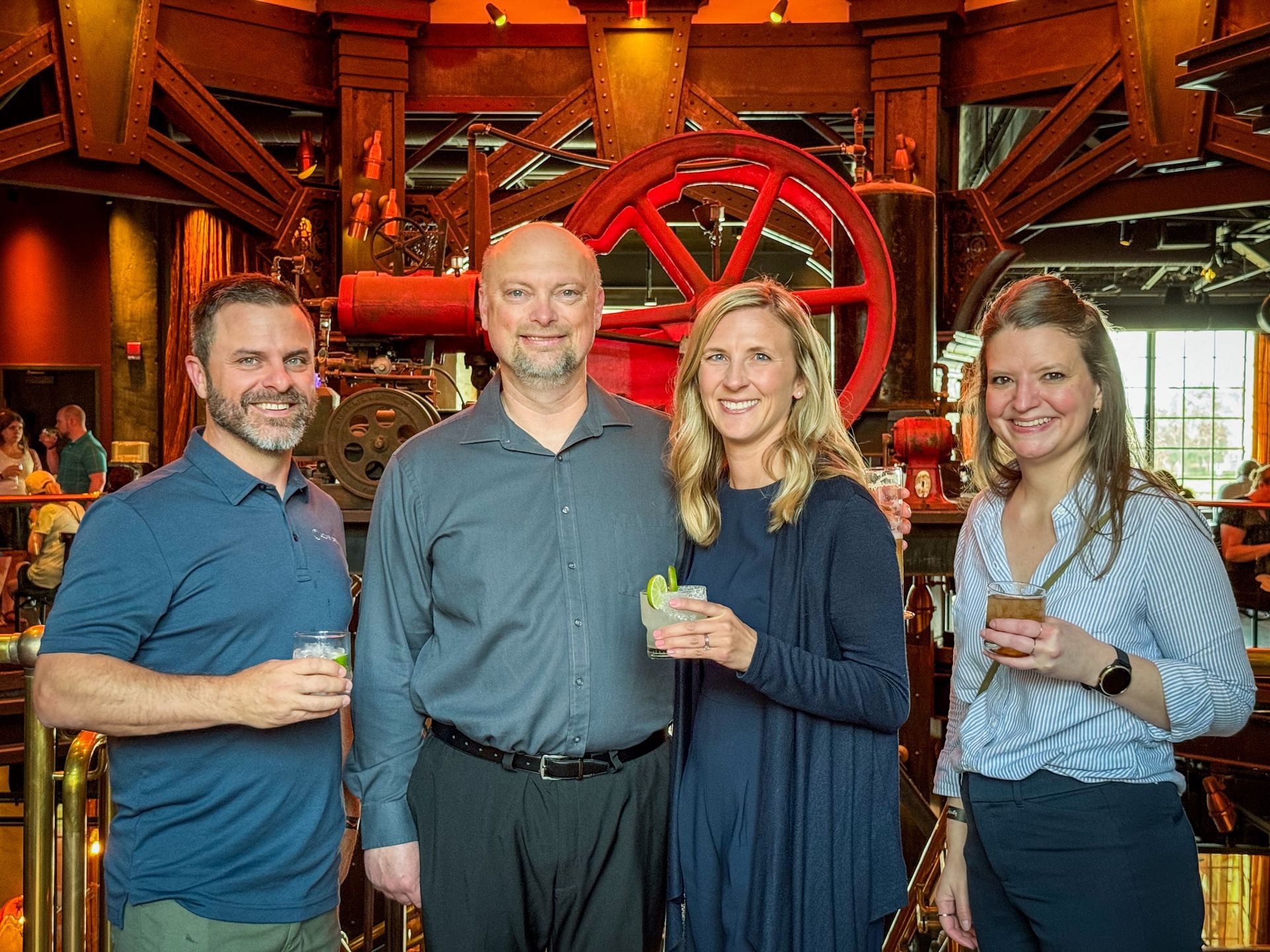 Four people smiling, holding drinks, standing in front of industrial machinery in a bar.