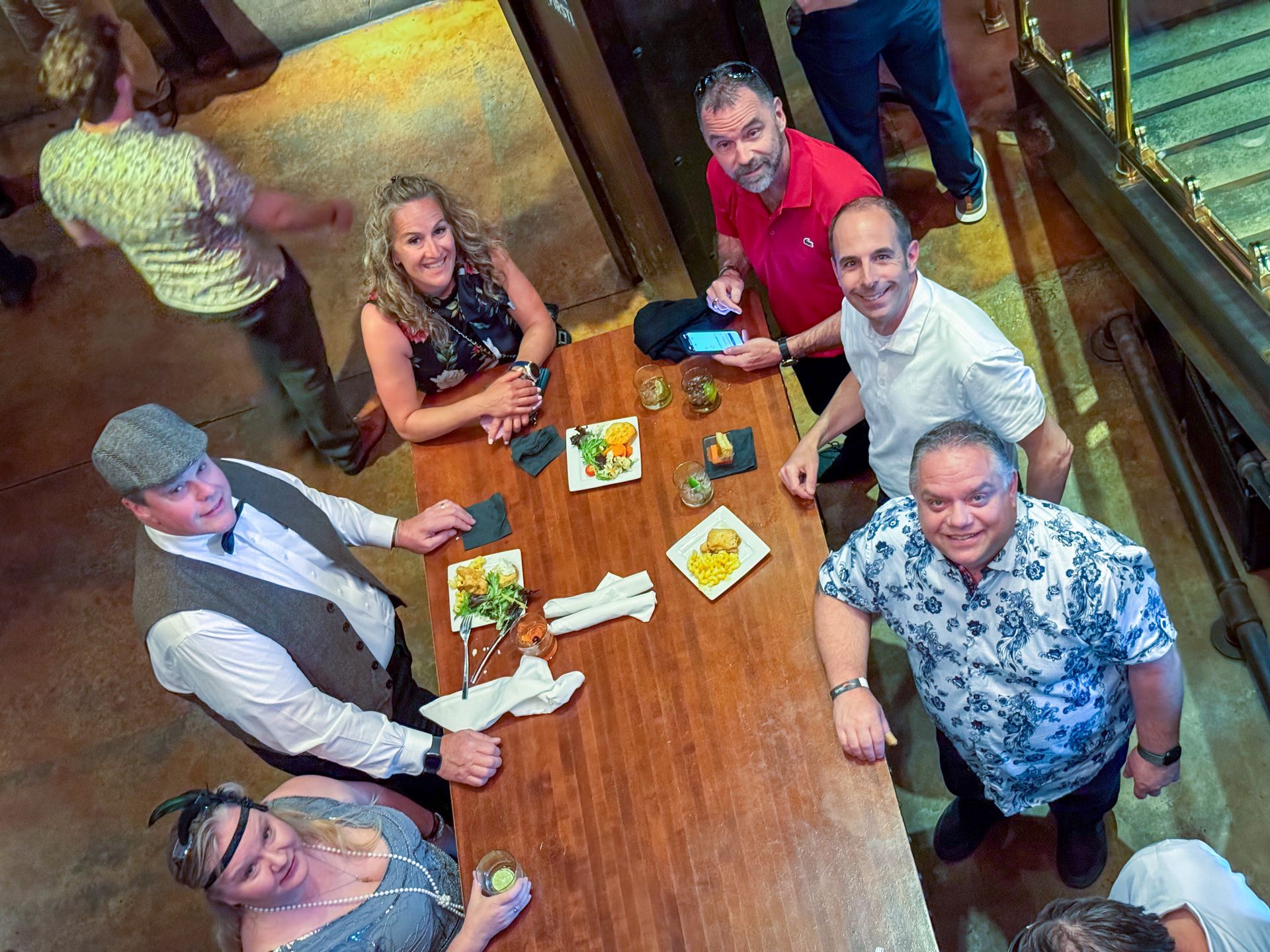 People gather around a wooden table in a dimly lit setting, some holding food and drinks.