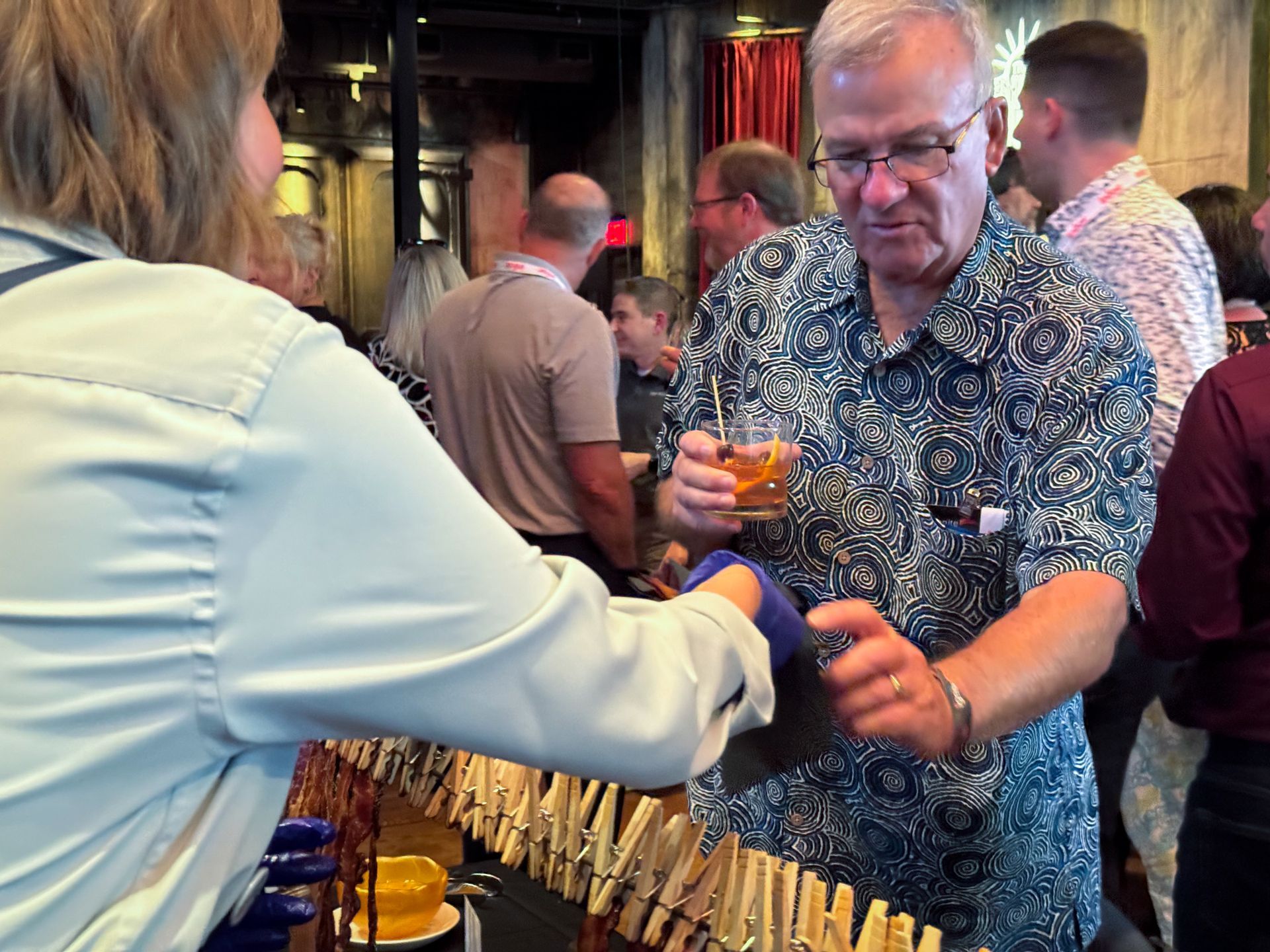 Man with drink being served at bar, wearing patterned shirt, interacting with server at a crowded event.