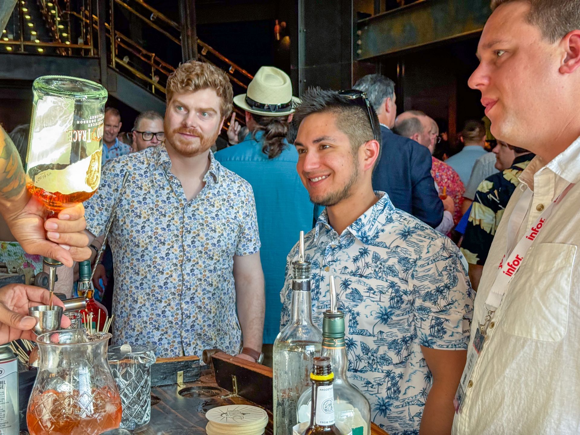 Men watching a bartender pouring a drink at a bar; bottles in front.