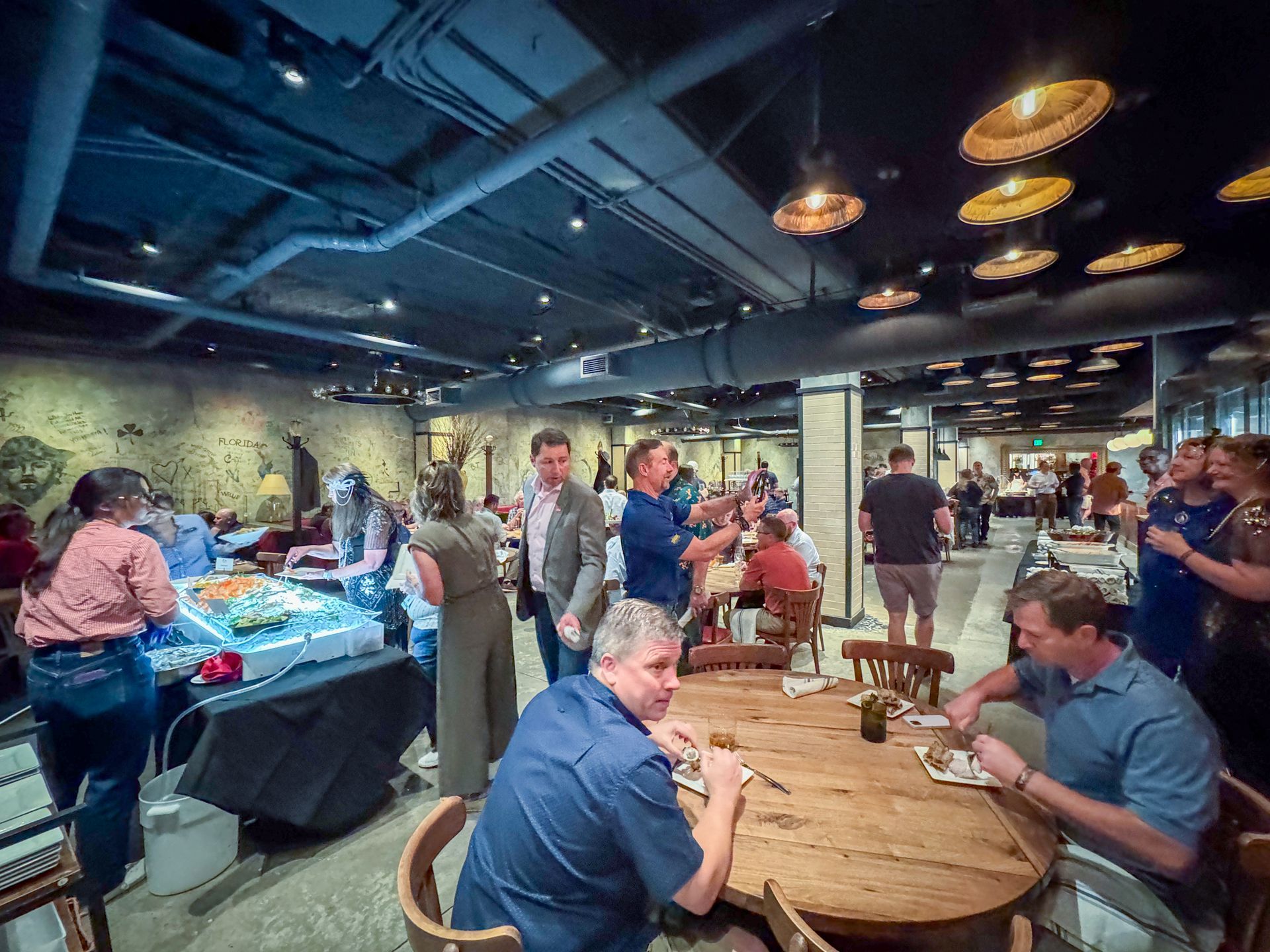 People socializing in a warmly lit event space with food tables, a mural, and decorative ceiling lights.
