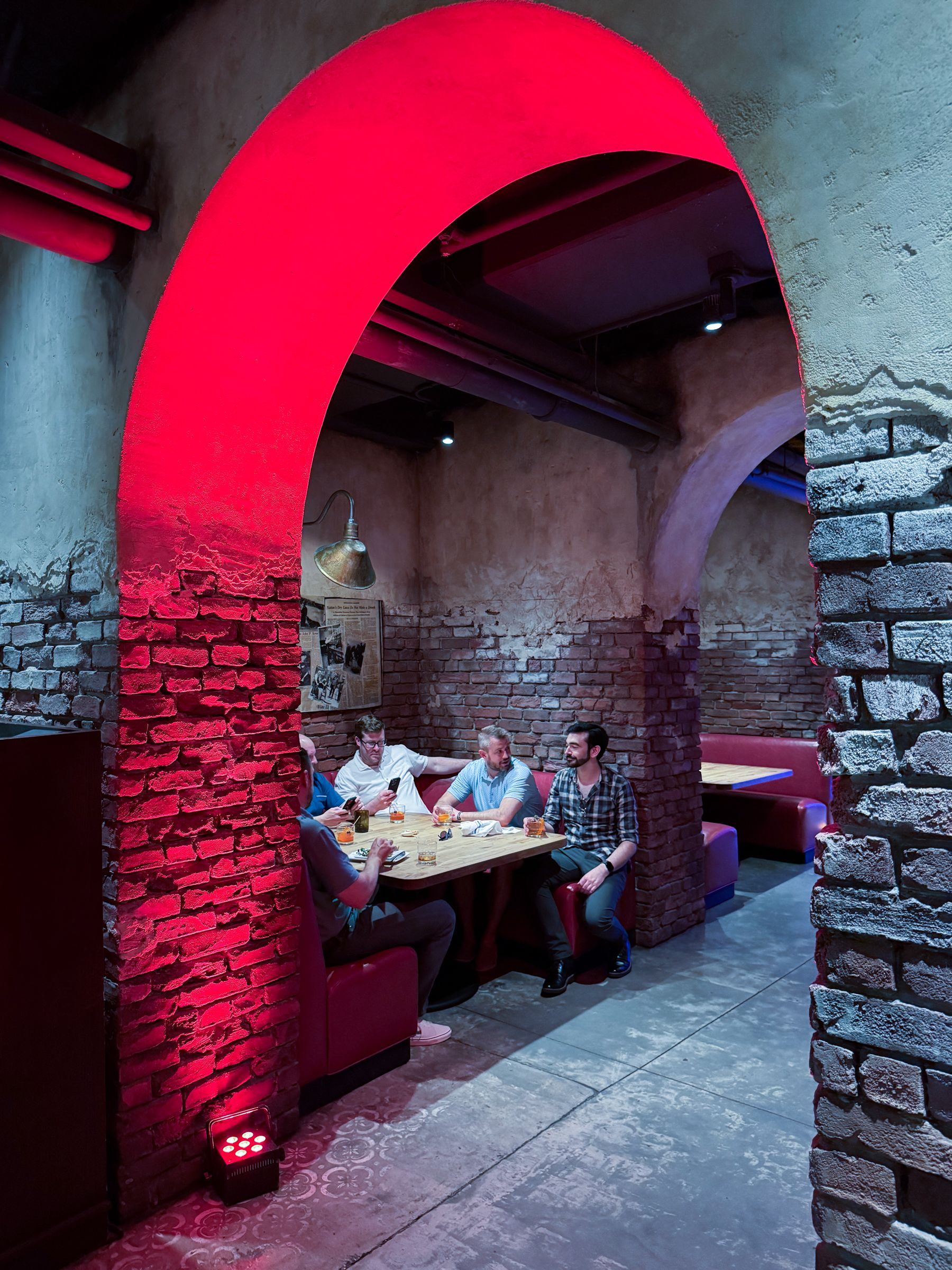 Red-lit archway leads to a restaurant dining area. Four men sit at a table. Brick walls and red booth seating are visible.