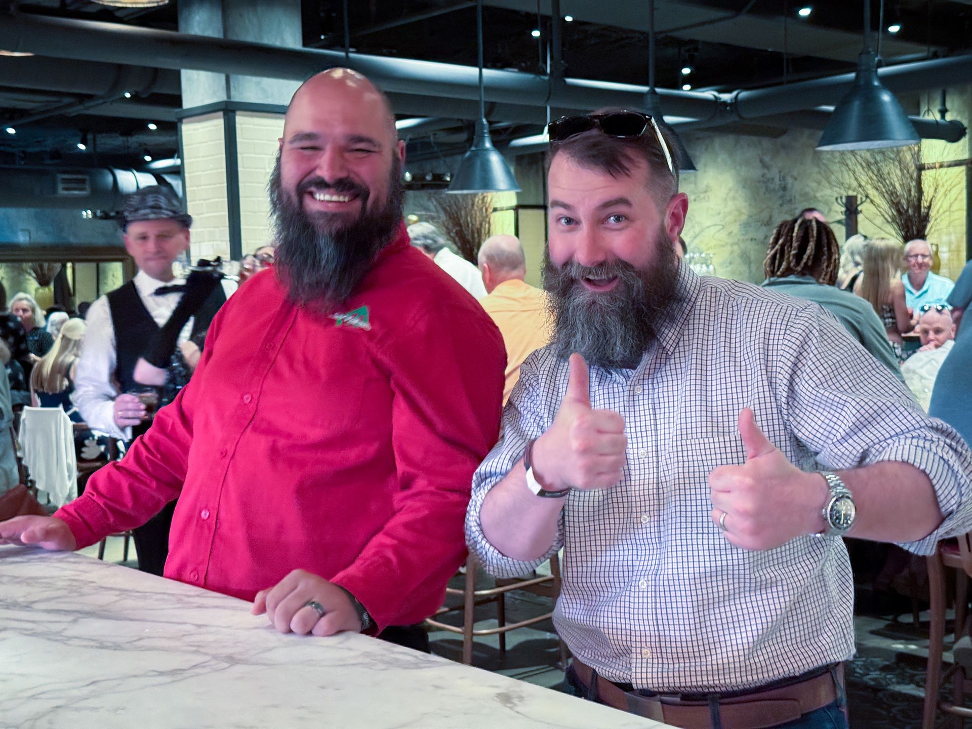 Two men with beards smile and give thumbs up at a bar. One in red, the other in a plaid shirt.