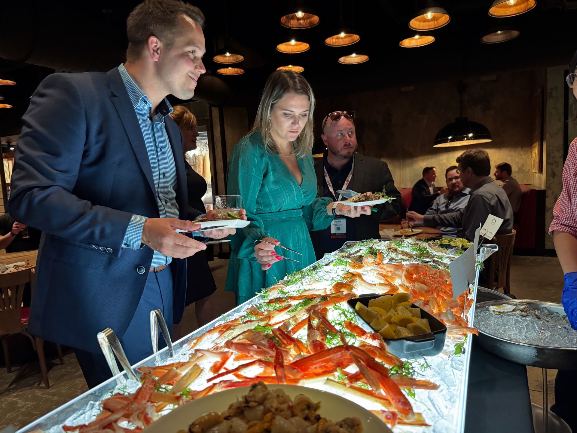 People at a buffet, selecting food. Seafood and sides are visible. Interior setting with hanging lights.