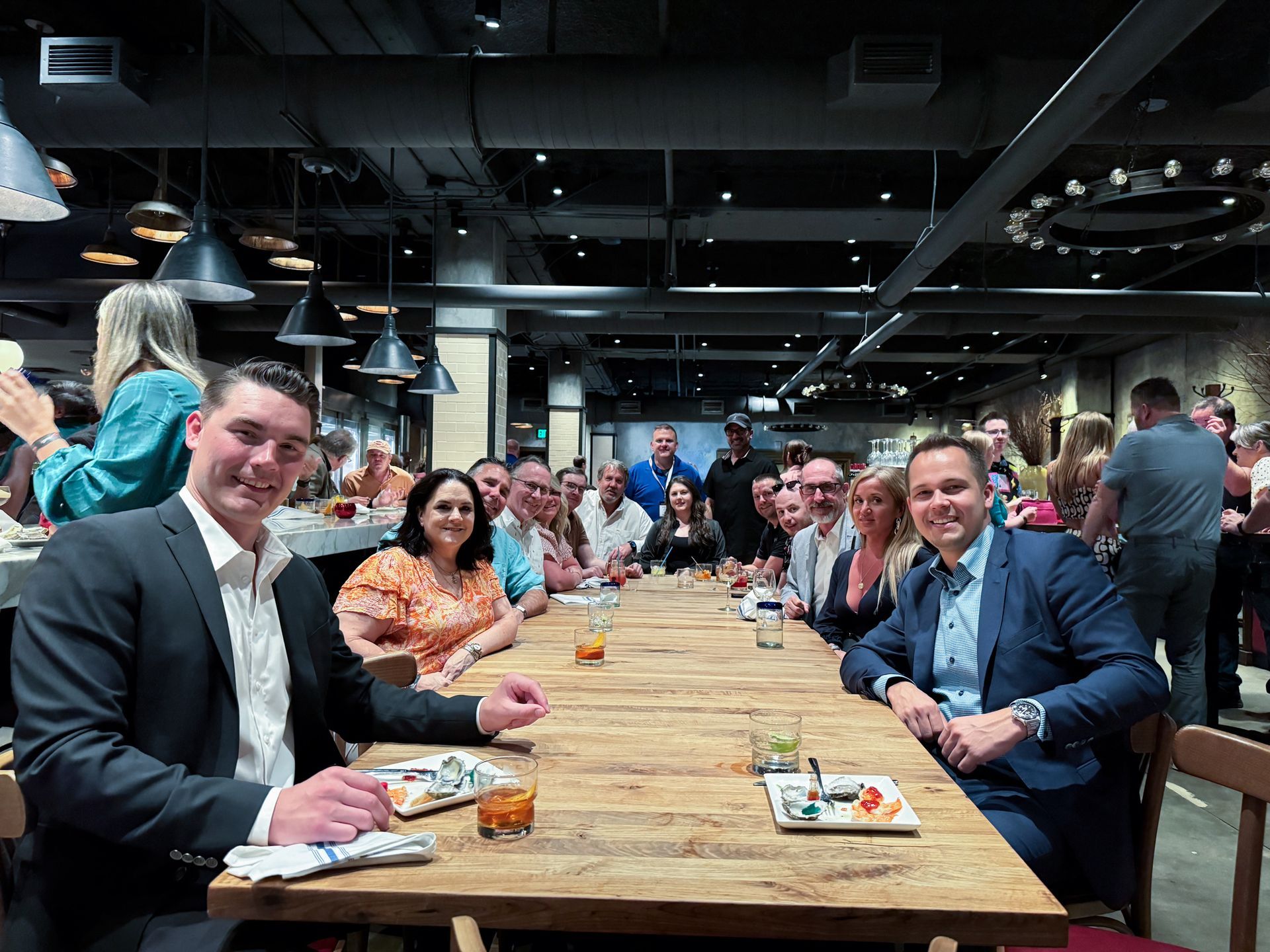 Group of people seated at a long wooden table in a restaurant; some smiling, some looking at camera.