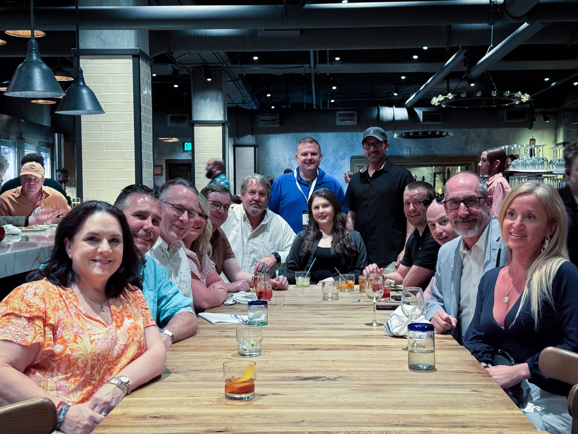 Group of people seated at a long wooden table in a restaurant. Some smiling, drinks present.