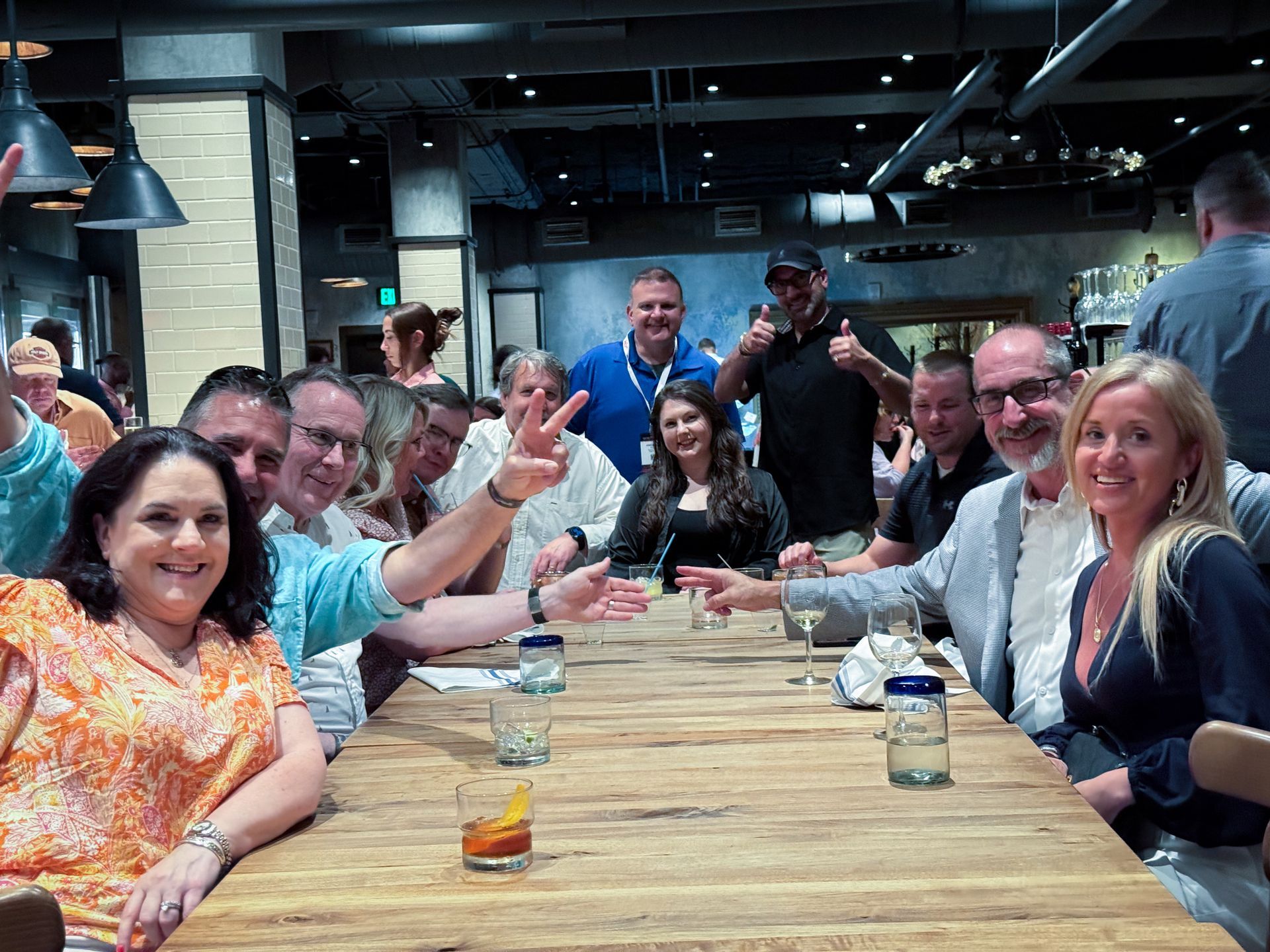 A group of people sitting at a long wooden table, some smiling and gesturing in a restaurant.