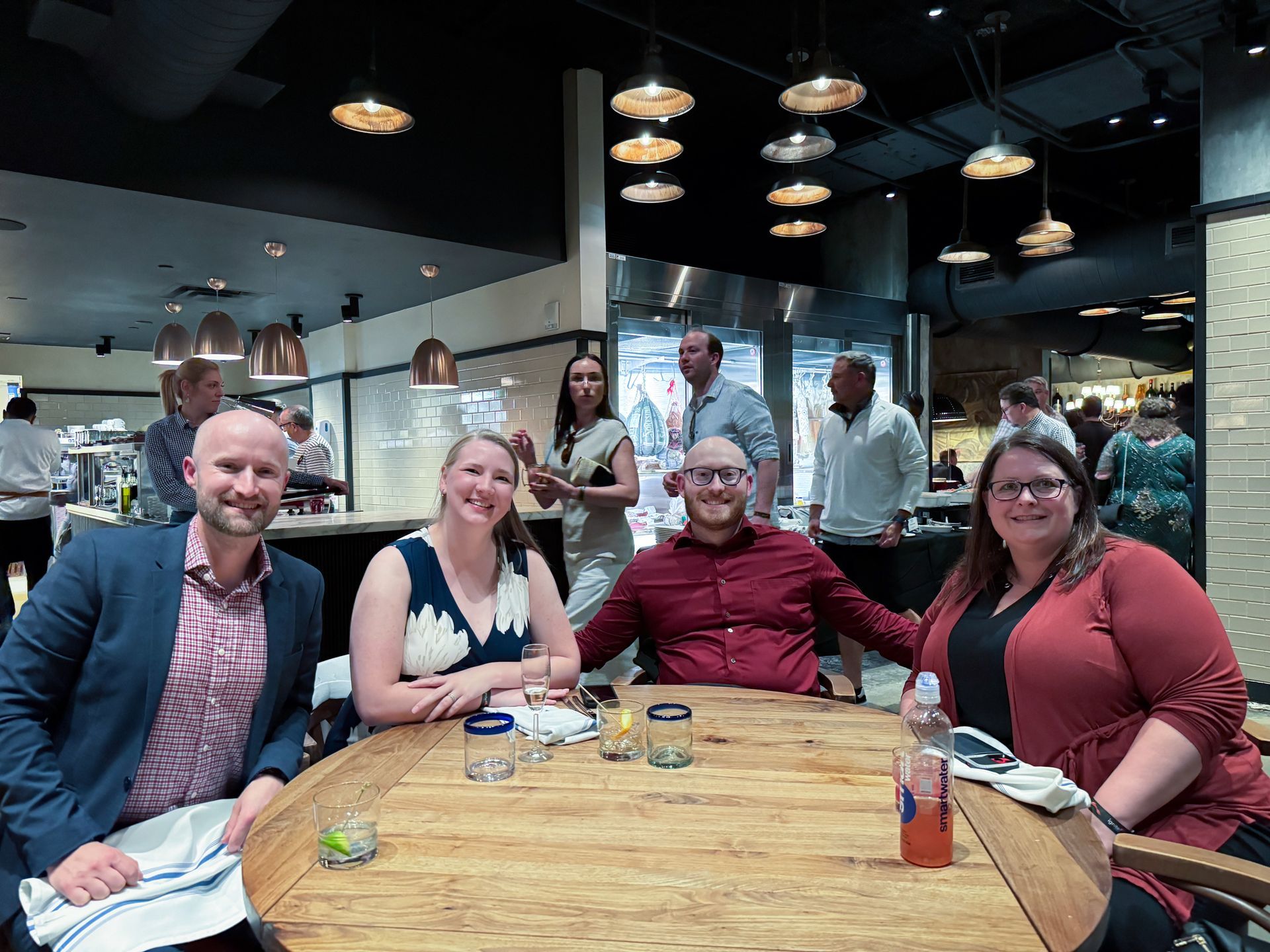 Four people seated at a wooden table in a restaurant. Others stand in the background.