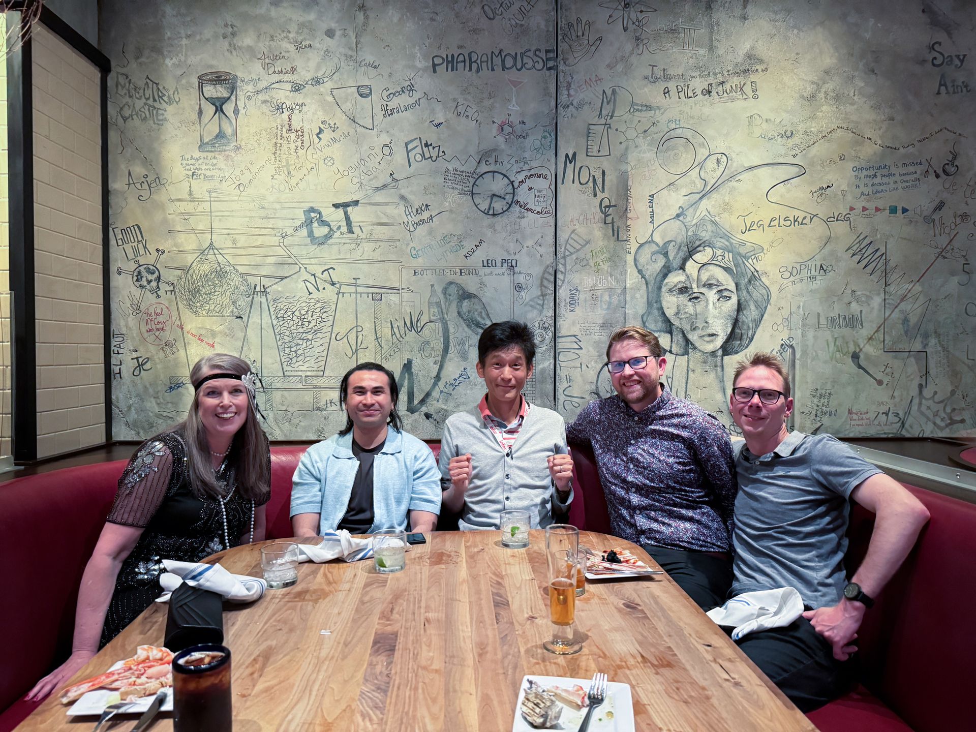 Five people seated around a wooden table in a booth with a graffiti-covered wall, smiling and posing.