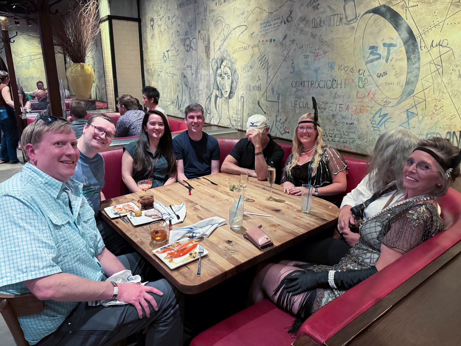 Group of people seated at a booth in a restaurant. Some are wearing 1920s attire. Graffiti on the wall.