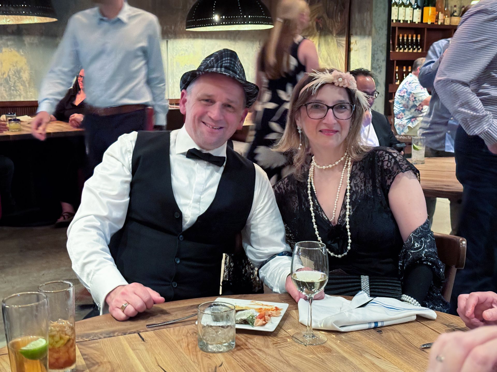 Couple in 1920s attire at a restaurant, smiling, sitting at a wooden table with drinks and food.