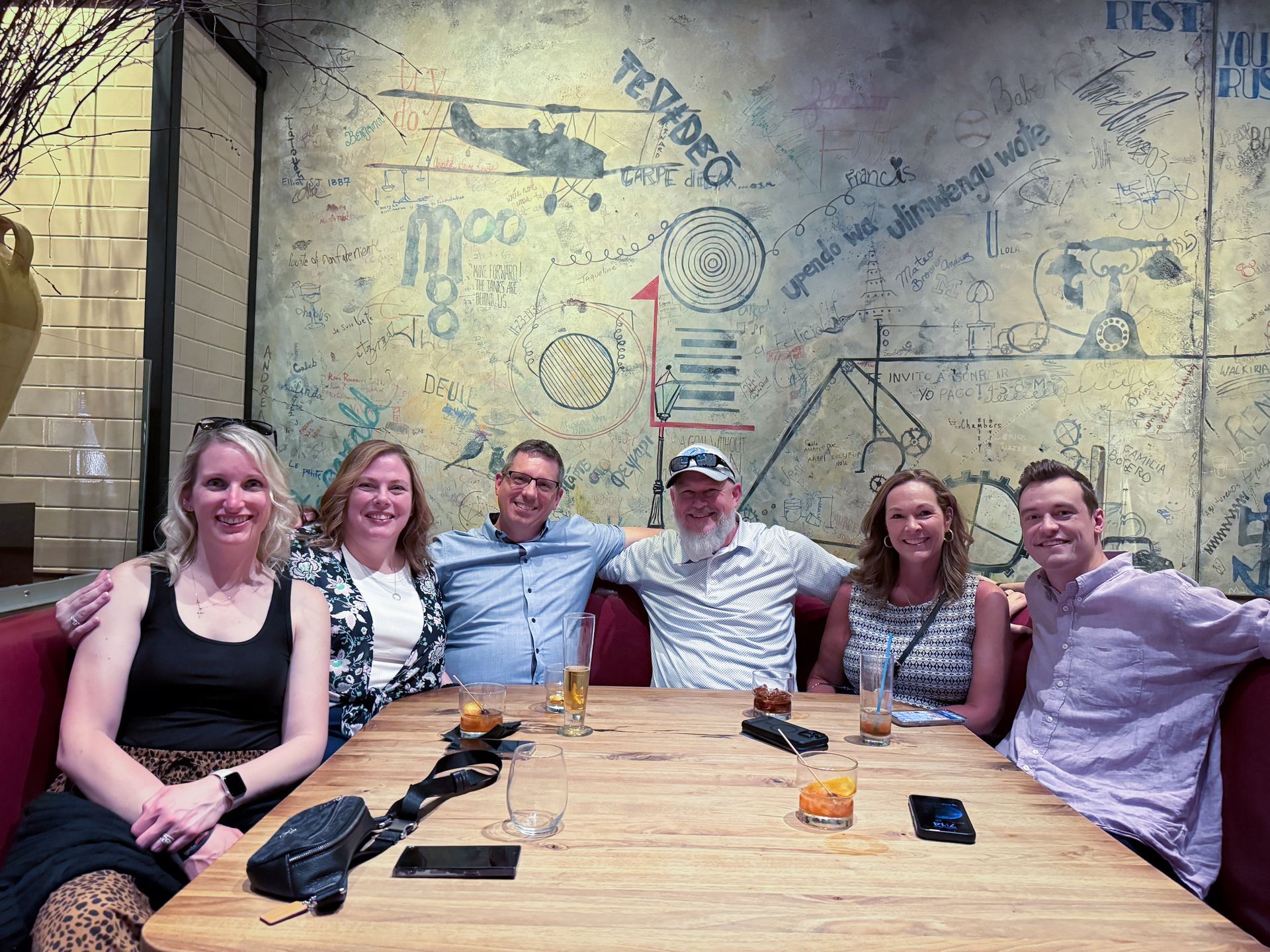 Six people seated at a table in a restaurant, arms around each other, smiles. Textured wall backdrop.