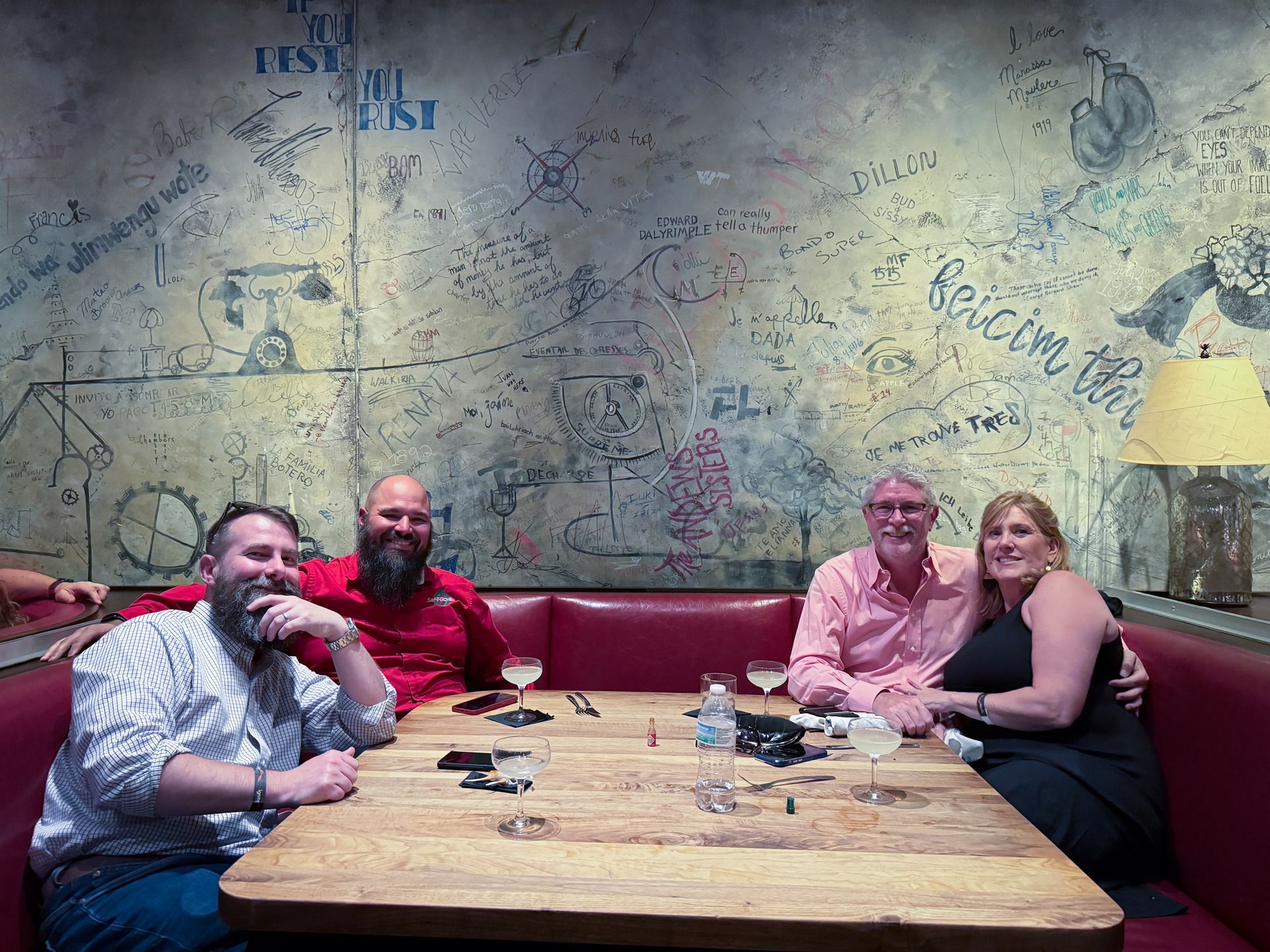 Four people seated at a table in a booth with graffiti on the wall. They are smiling, with drinks.
