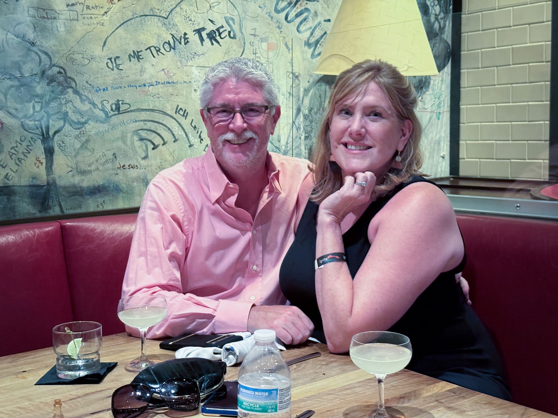 Couple seated at table, smiling. Man in pink shirt, woman in black dress. Drinks on the table, interior setting.