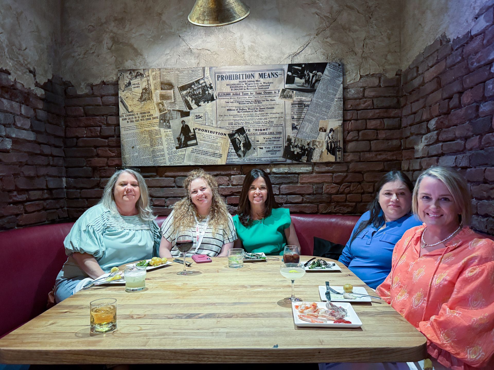 Five women seated at a table in a restaurant, eating and smiling. Brick wall and abstract art in background.