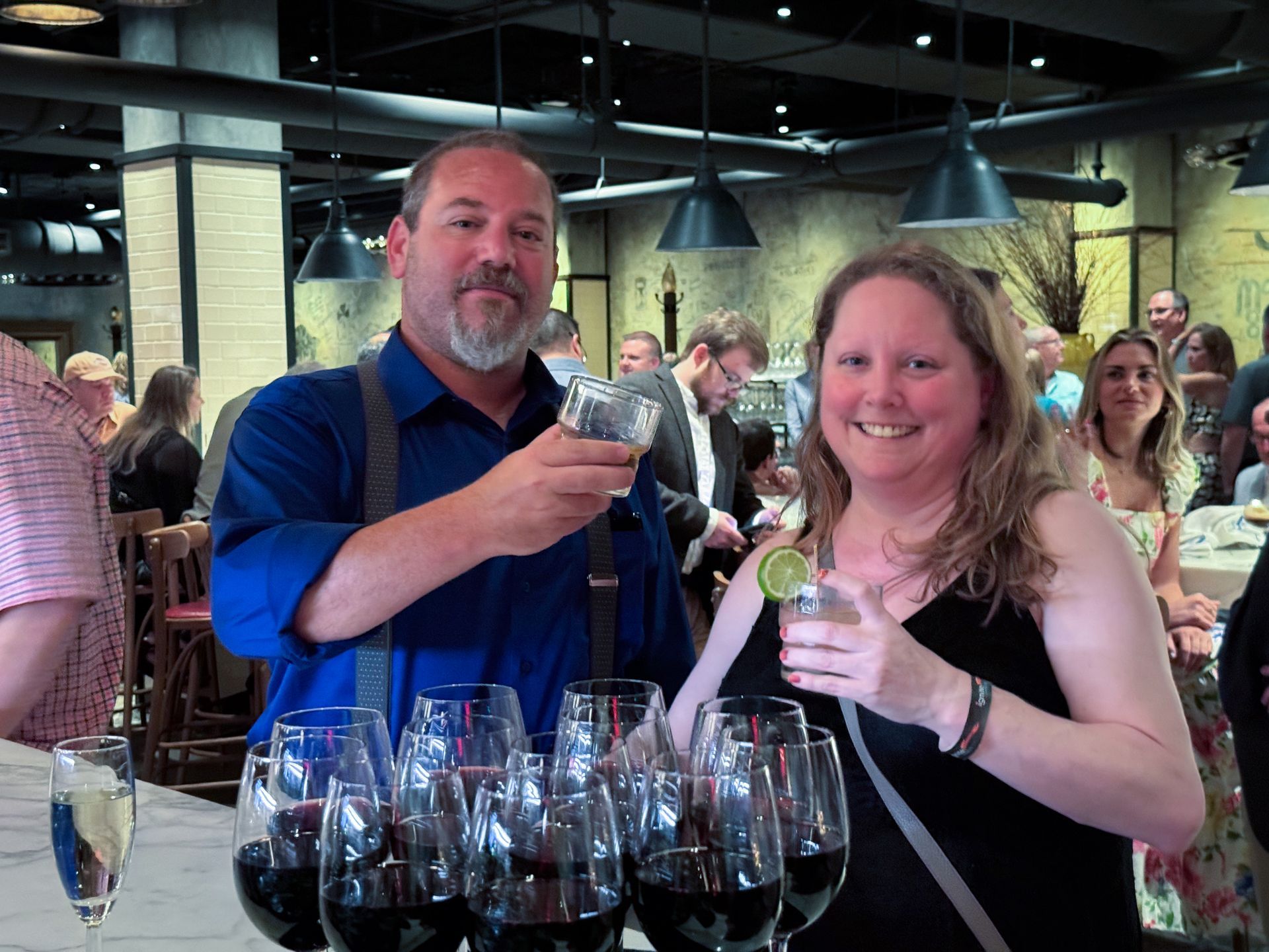 Man and woman toasting drinks in a bar. Many wine glasses on counter. Others in background.