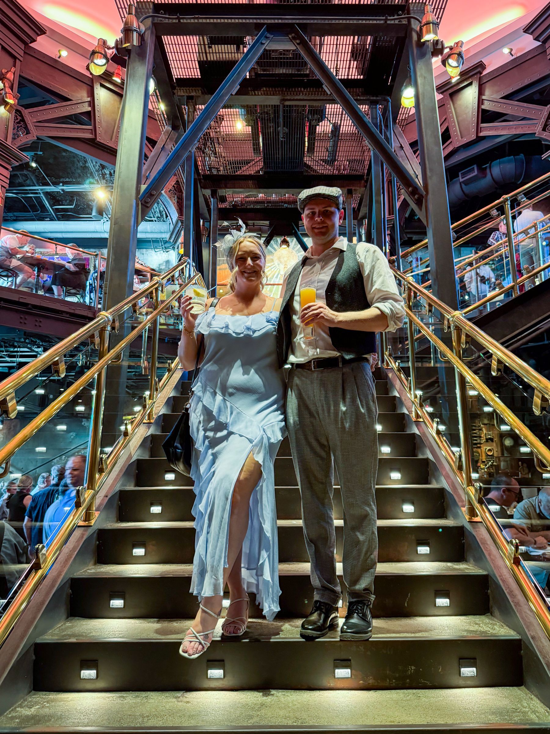 Woman in blue dress and man in vest stand on ornate staircase. They are smiling and holding drinks.