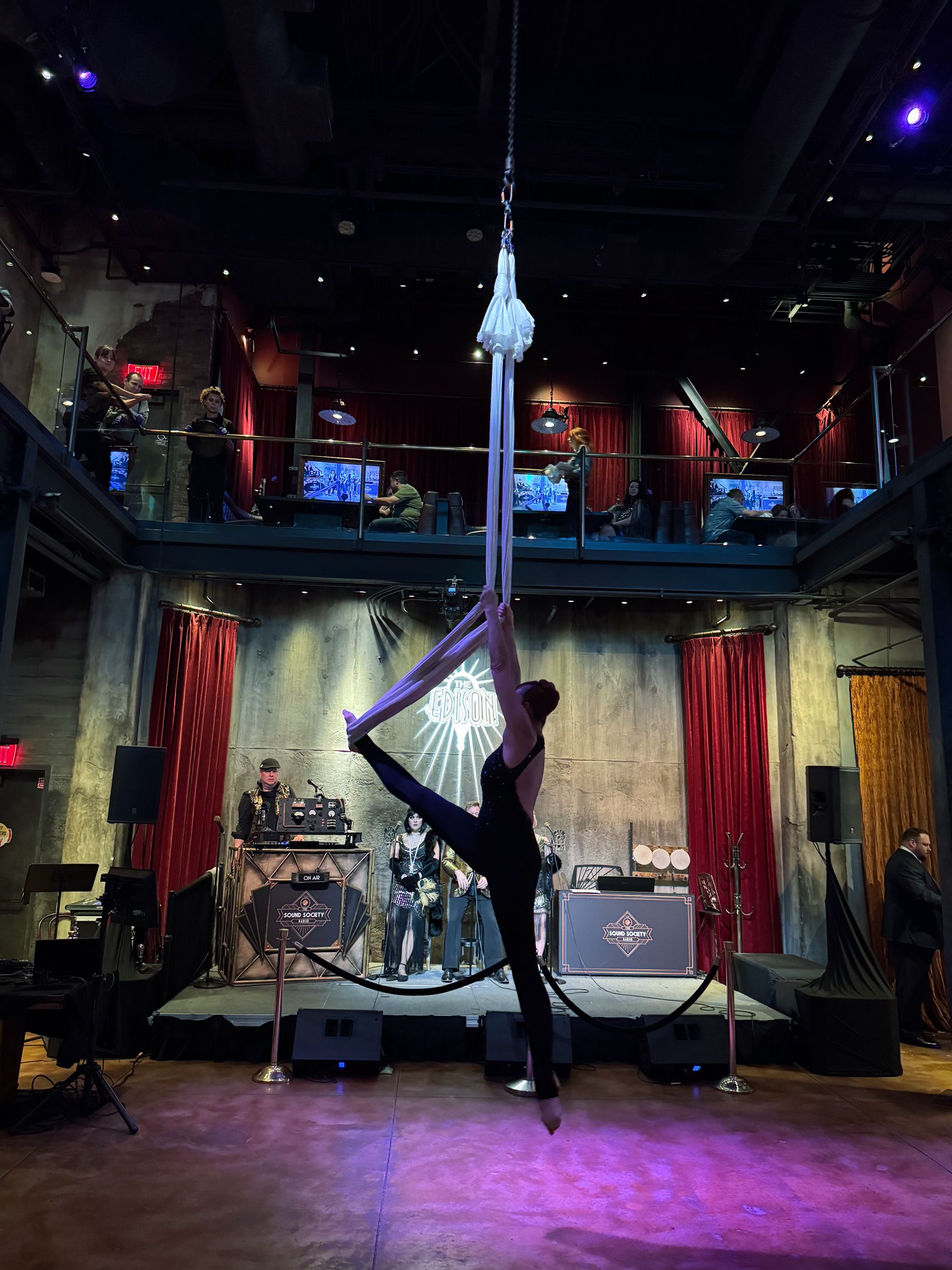 Aerial performer doing a split on a silk apparatus, in a dimly lit venue with a stage and audience.