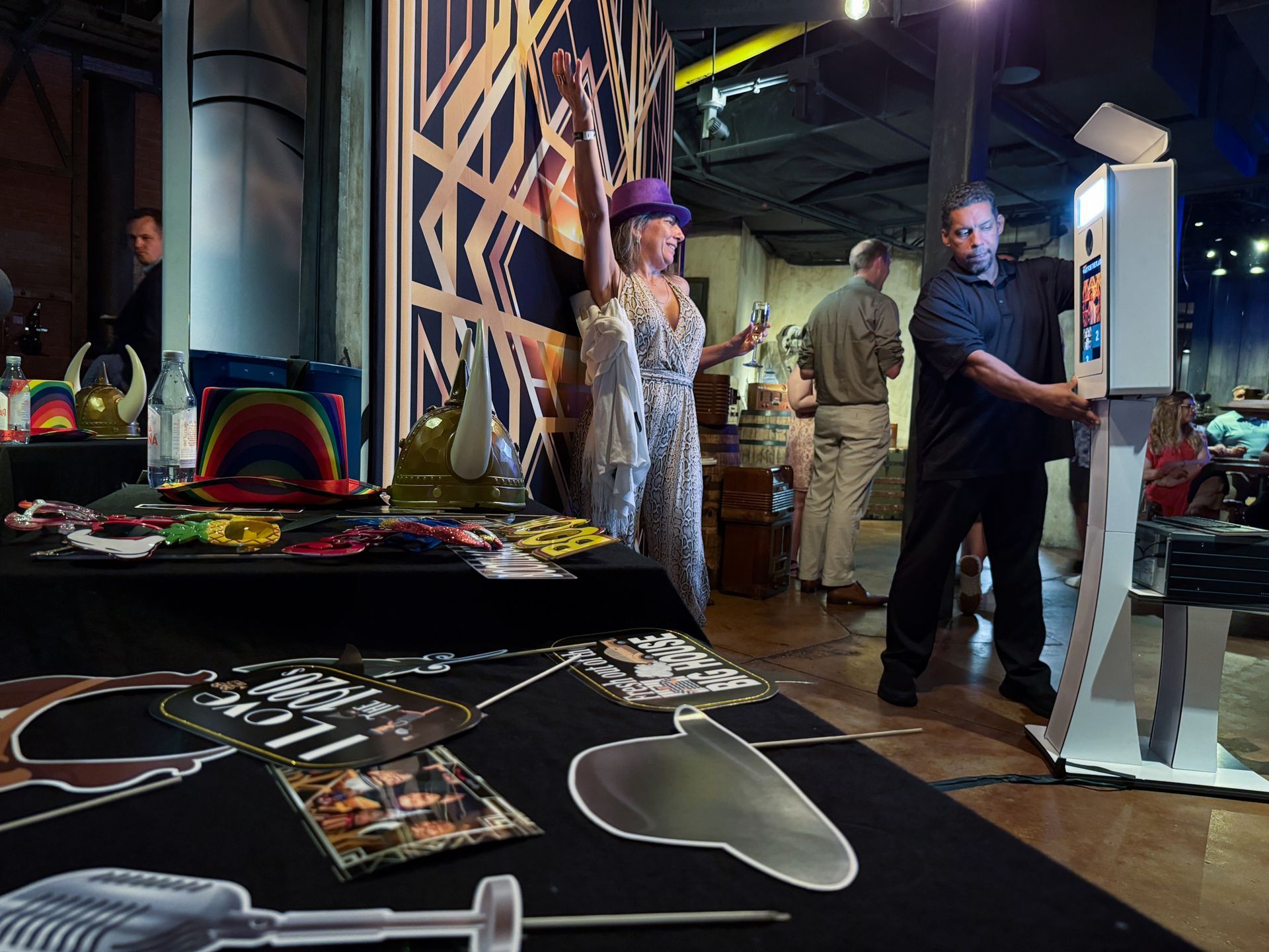 People at event with photo booth. Woman in sparkly outfit poses. Man uses booth. Props on table.